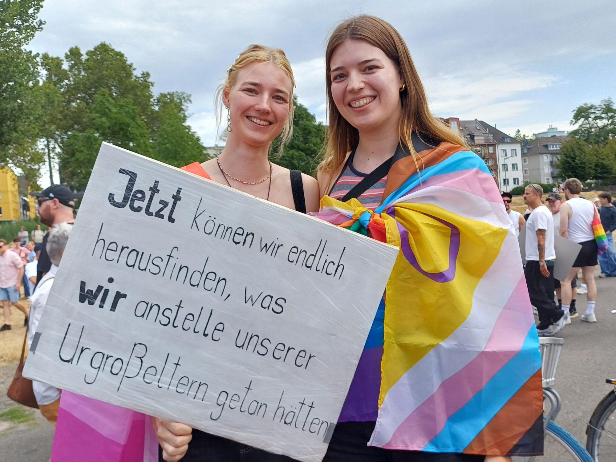 Zwei junge Frauen halten ein Plakat mit der Aufschrift: Jetzt können wir endlich herausfinden, was wir anstelle unserer Urgroßeltern getan hätten.