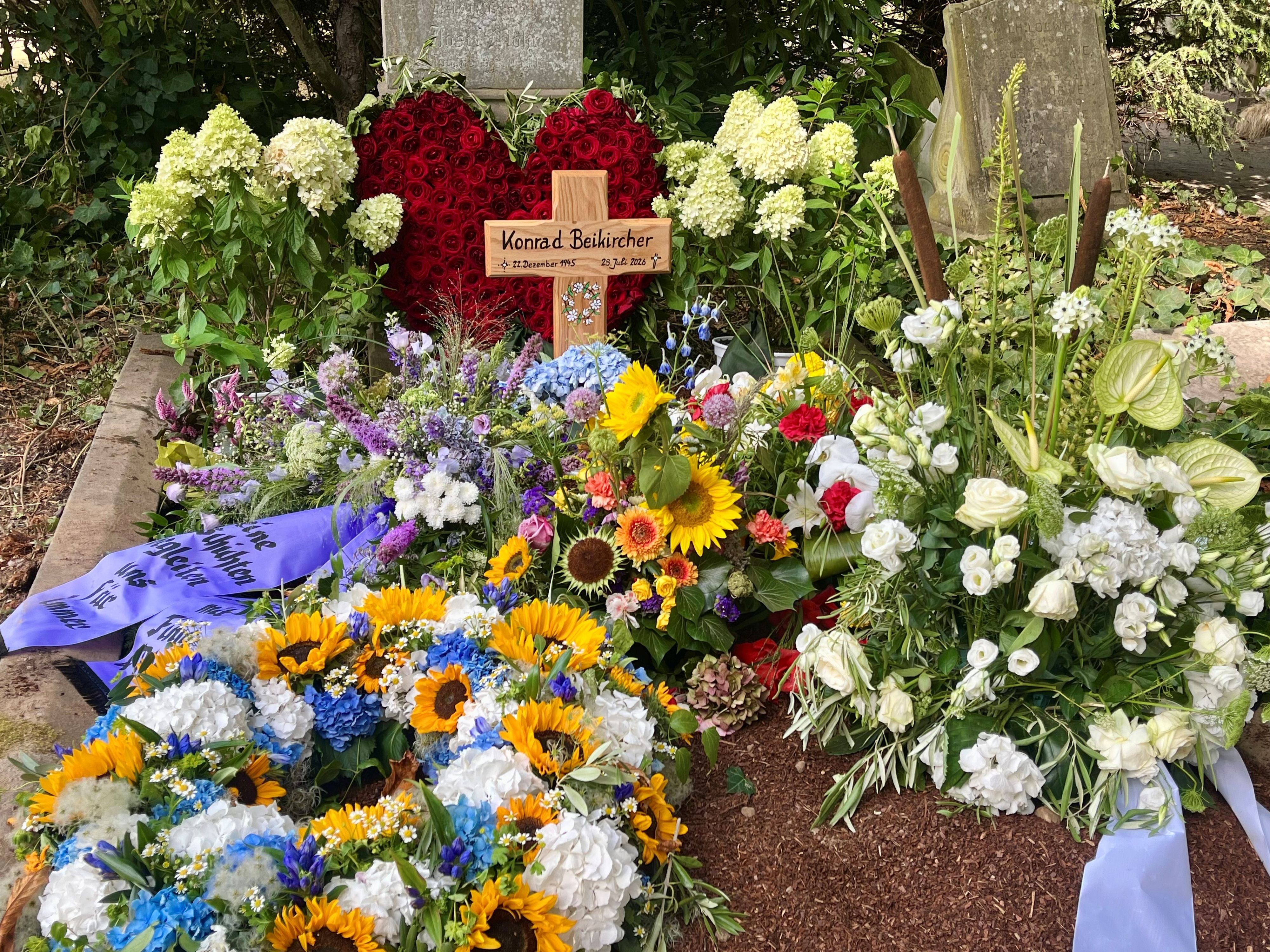 Auf dem Grab von Konrad Beikirchen steht ein Holzkreuz mit seinem Namen und seinen Lebensdaten, liegen mehrere Blumenkränze sowie ein Herz aus roten Rosen.