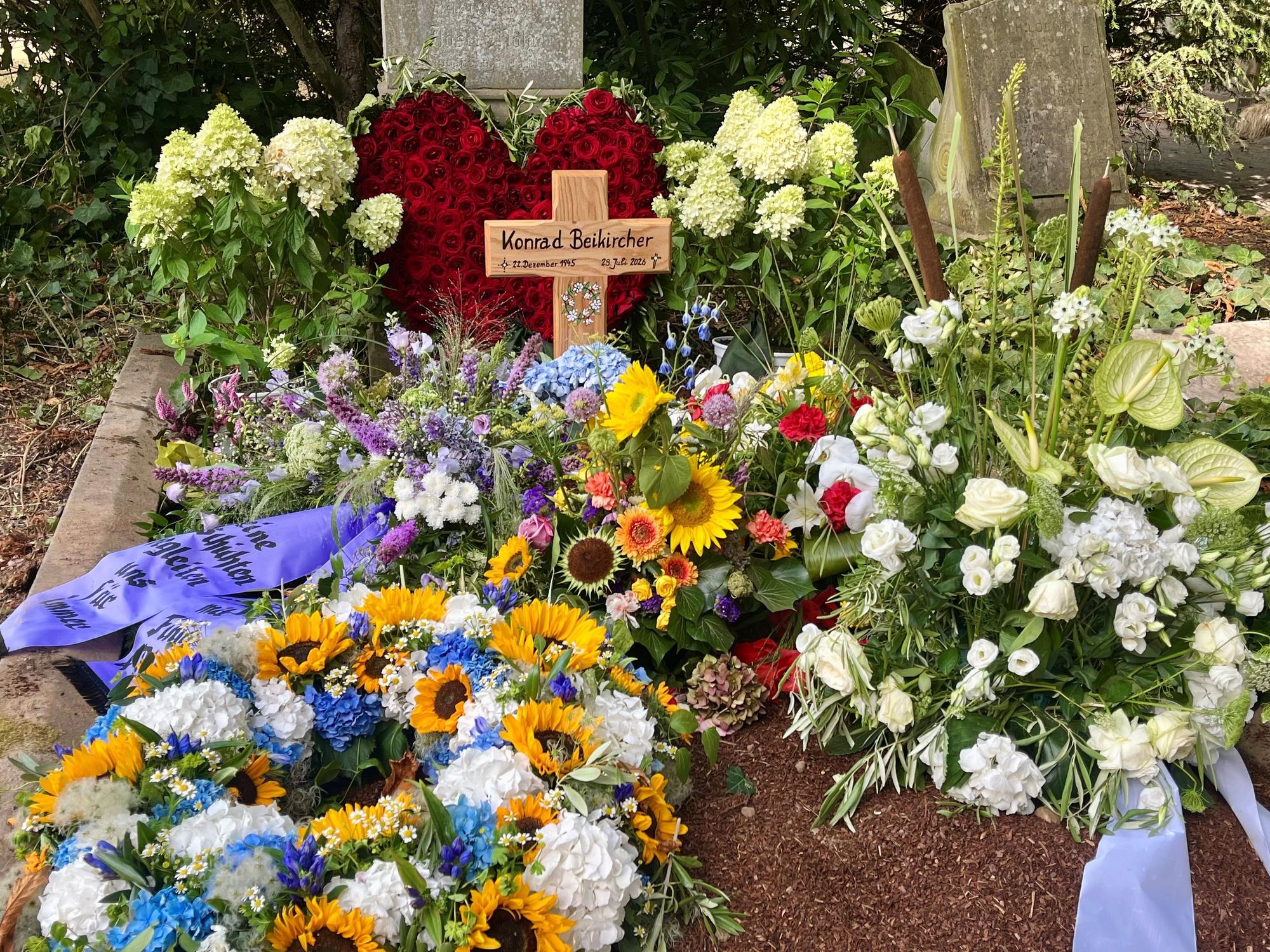 Auf dem Grab von Konrad Beikirchen steht ein Holzkreuz mit seinem Namen und seinen Lebensdaten, liegen mehrere Blumenkränze sowie ein Herz aus roten Rosen.
