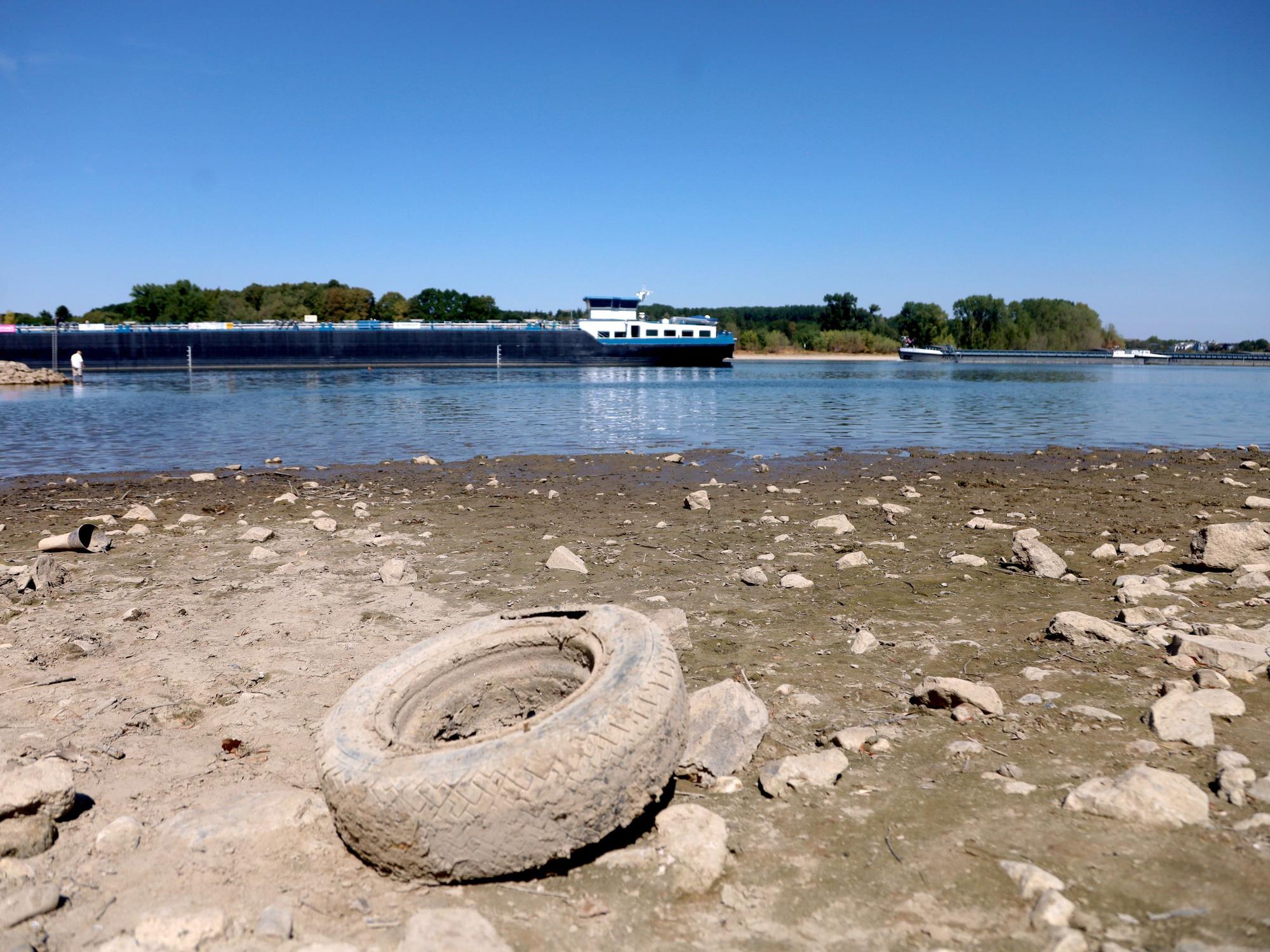 Der sinkende Rheinpegel hat einen Autoreifen trockengelegt, im Hintergrund ist ein Schiff im Rhein unterwegs.