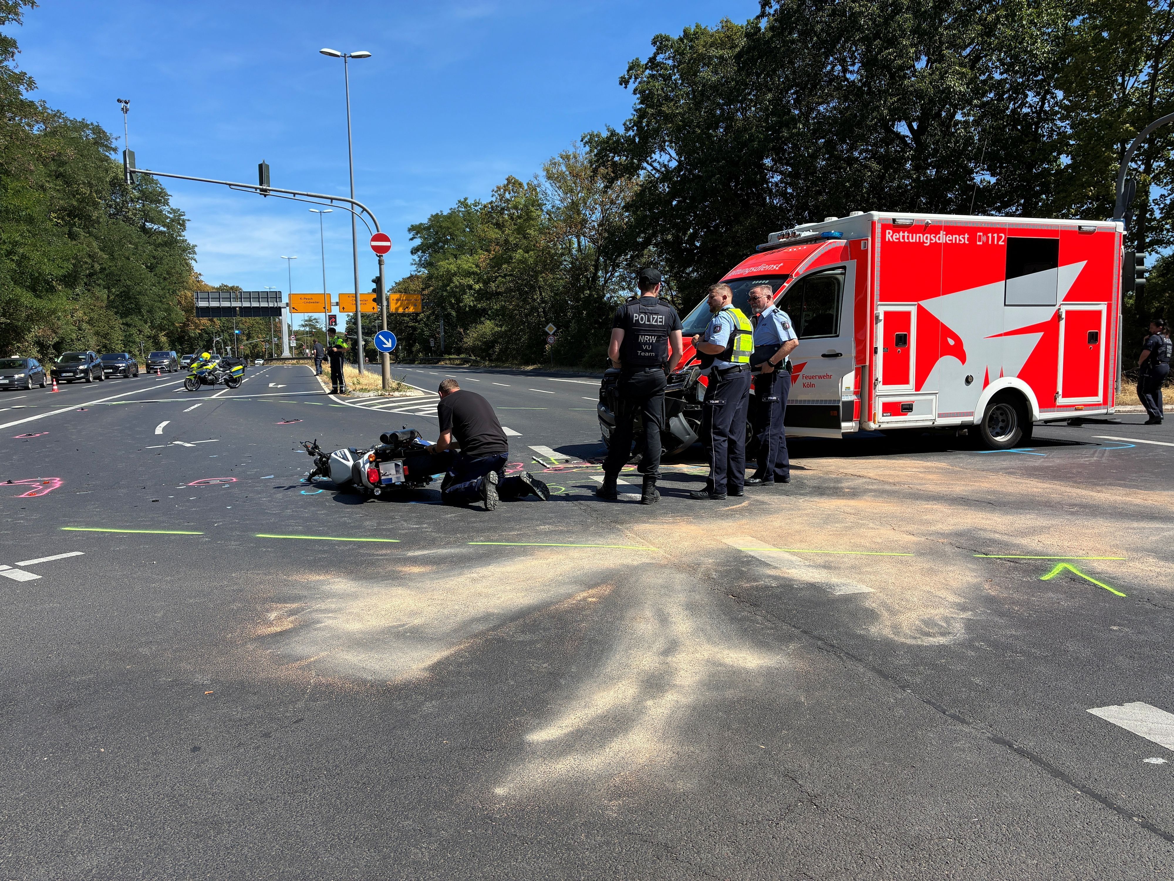 Vor einem RTW liegt ein Motorrad auf der Fahrbahn, Polizisten stehen davor, einer kniet.