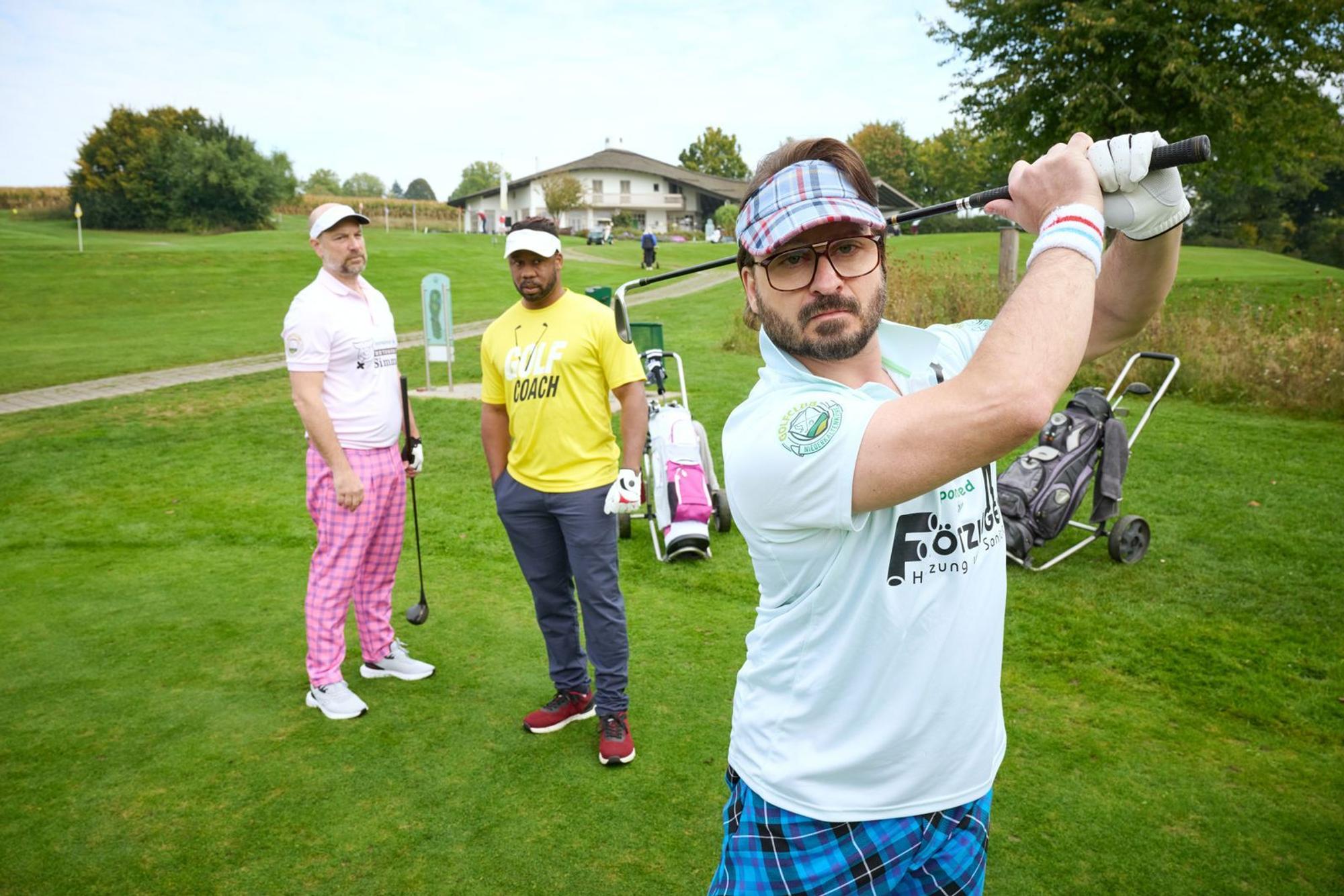 Simmerl (Stephan Zinner, l.) und Flötzinger (Daniel Christensen, r.) versuchen sich unter Aufsicht von Golflehrer Buengo (Castro Dokyi Affum) auf dem Grün. (Bild: Constantin Film Distribution GmbH / Bernd Schuller)