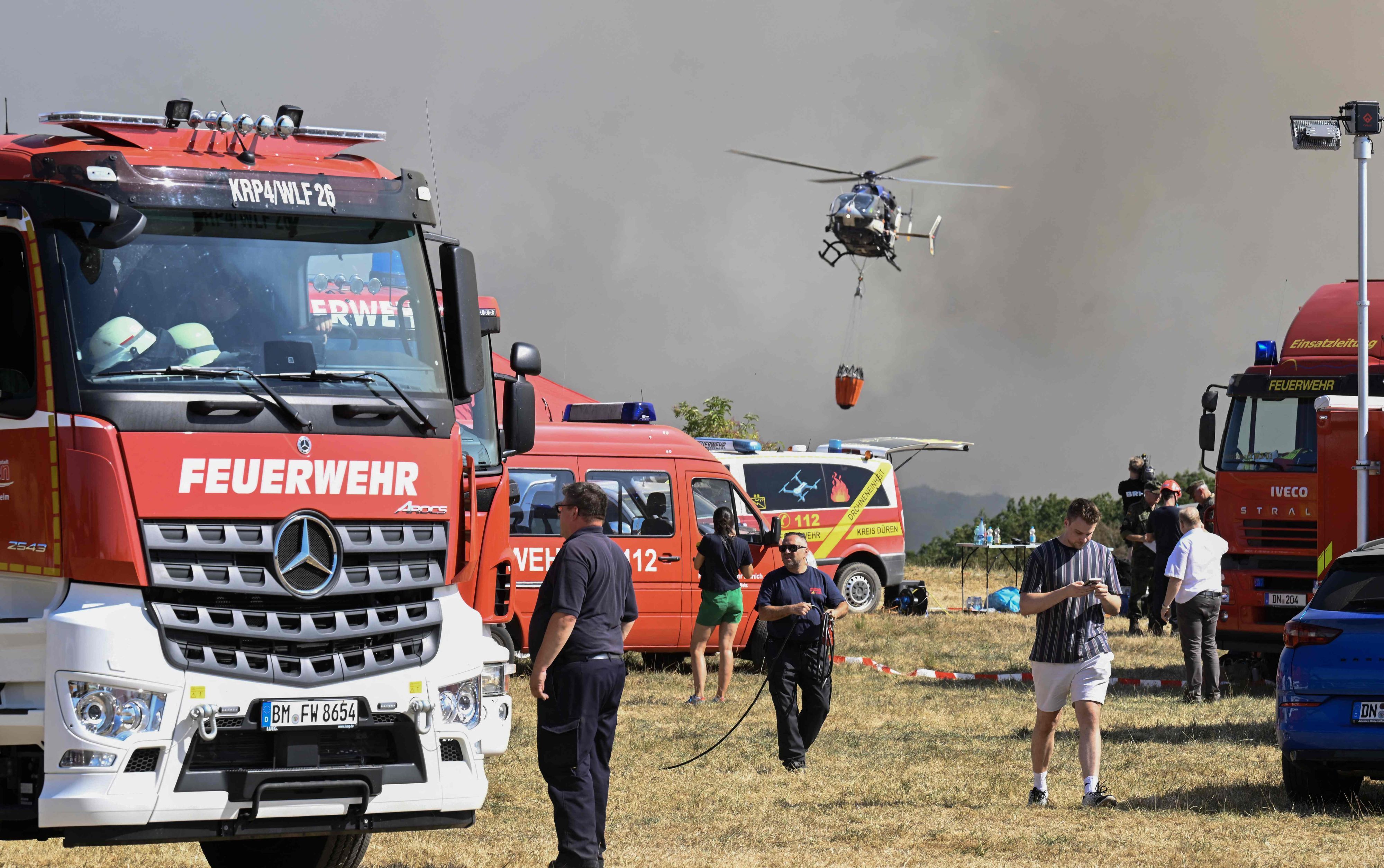Ein Hubschrauber ist vor einer Rauchwand in der Luft, auf dem Boden stehen zahlreiche Einsatzfahrzeuge der Feuerwehr.