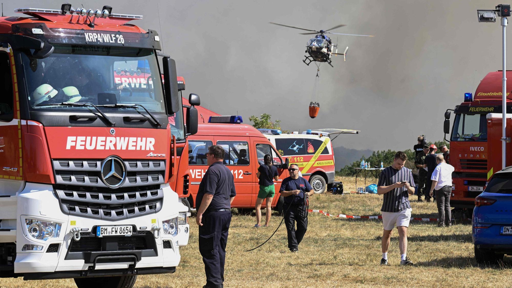 Ein Hubschrauber ist vor einer Rauchwand in der Luft, auf dem Boden stehen zahlreiche Einsatzfahrzeuge der Feuerwehr.