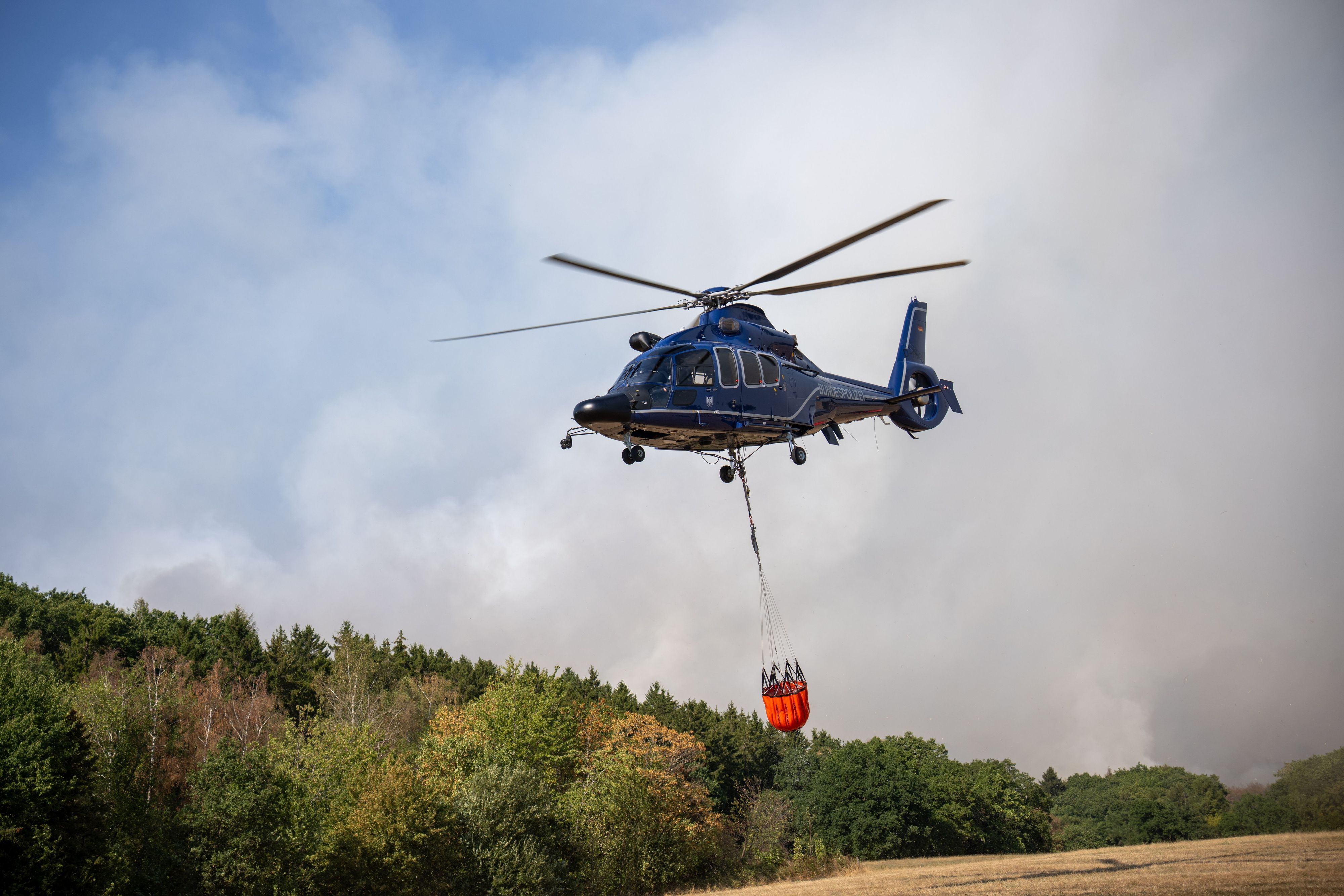 14.08.2026, Nordrhein-Westfalen, Hürtgenwald: Ein Helikopter der Bundespolizei mit einem Löschwasser-Außenlastbehälter ist beim Waldbrand im Hürtgenwald im Einsatz. Mehrere Hundert Hektar Waldfläche stehen in Flammen. (zu dpa: «Mehrere Löschhubschrauber bei Hürtgenwald erwartet») Foto: Harald Tittel/dpa +++ dpa-Bildfunk +++
