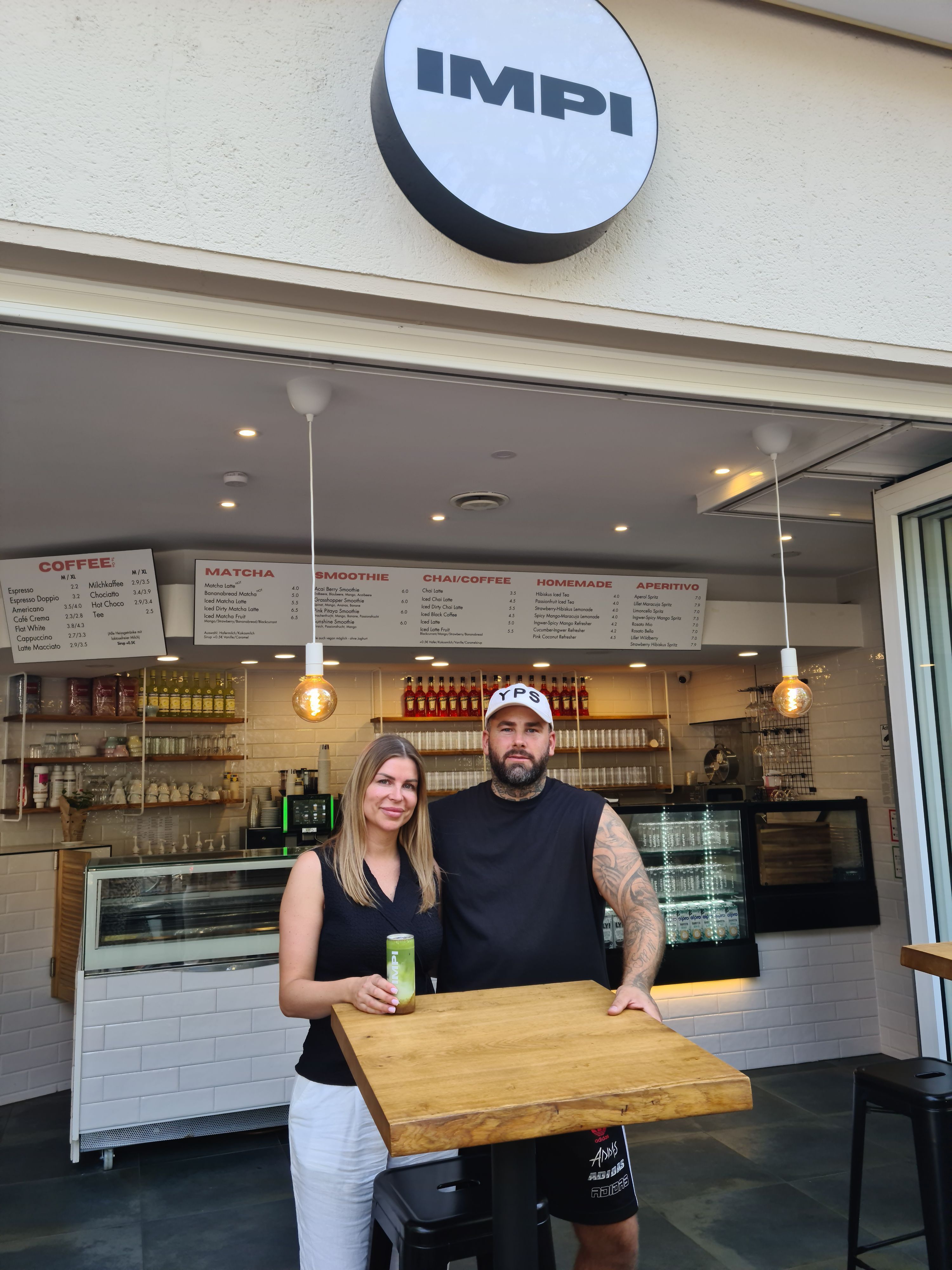 Eine Frau mit blondem Haar steht neben einem Mann mit Bart und Basecap hinter dem Stehtisch eines Cafés.