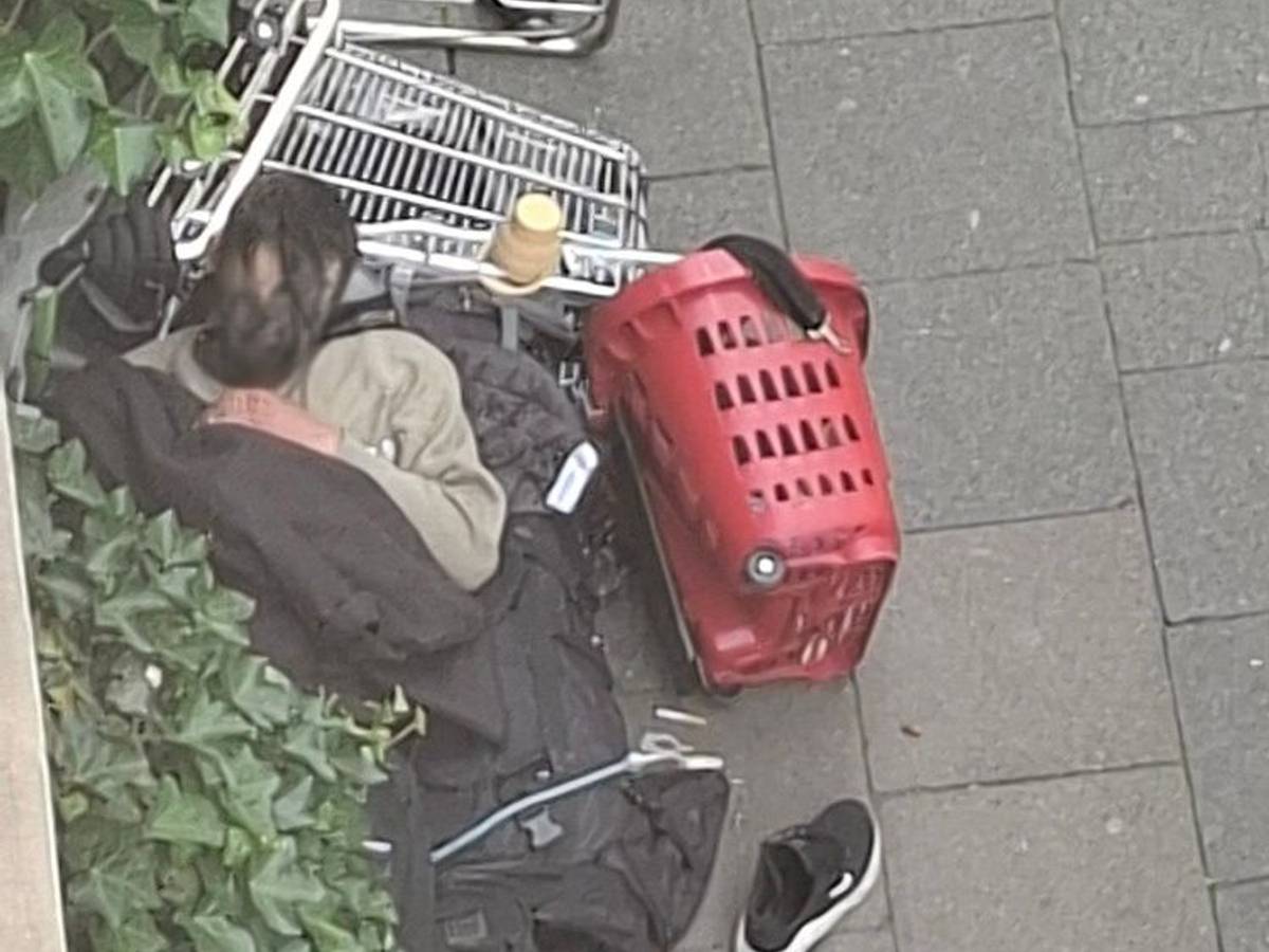 Ein Mann schläft auf dem Gehweg, den Kopf auf einem umgekippten Einkaufswagen.