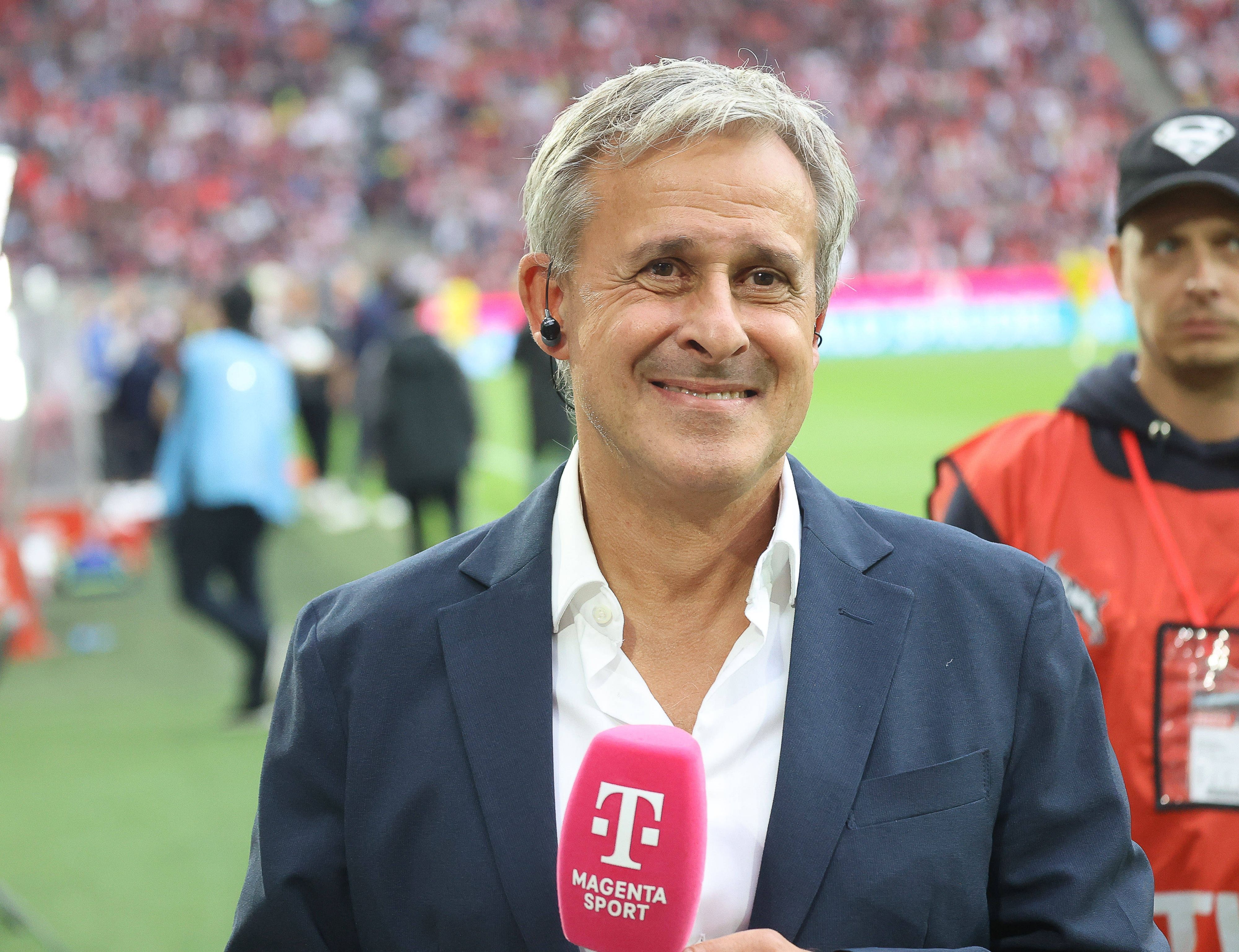 Pierre Littbarski steht mit Mikrofon im Kölner Stadion und grinst in die Kamera.