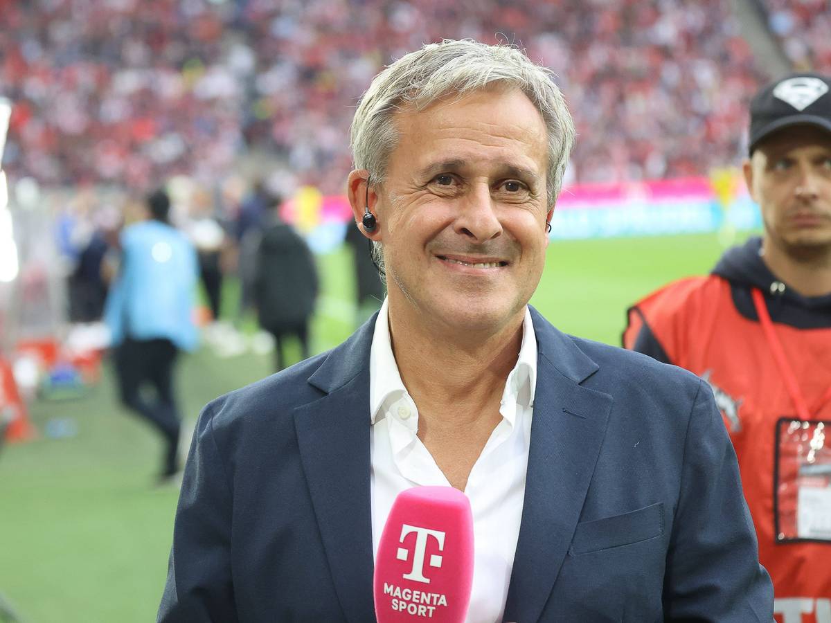 Pierre Littbarski steht mit Mikrofon im Kölner Stadion und grinst in die Kamera.