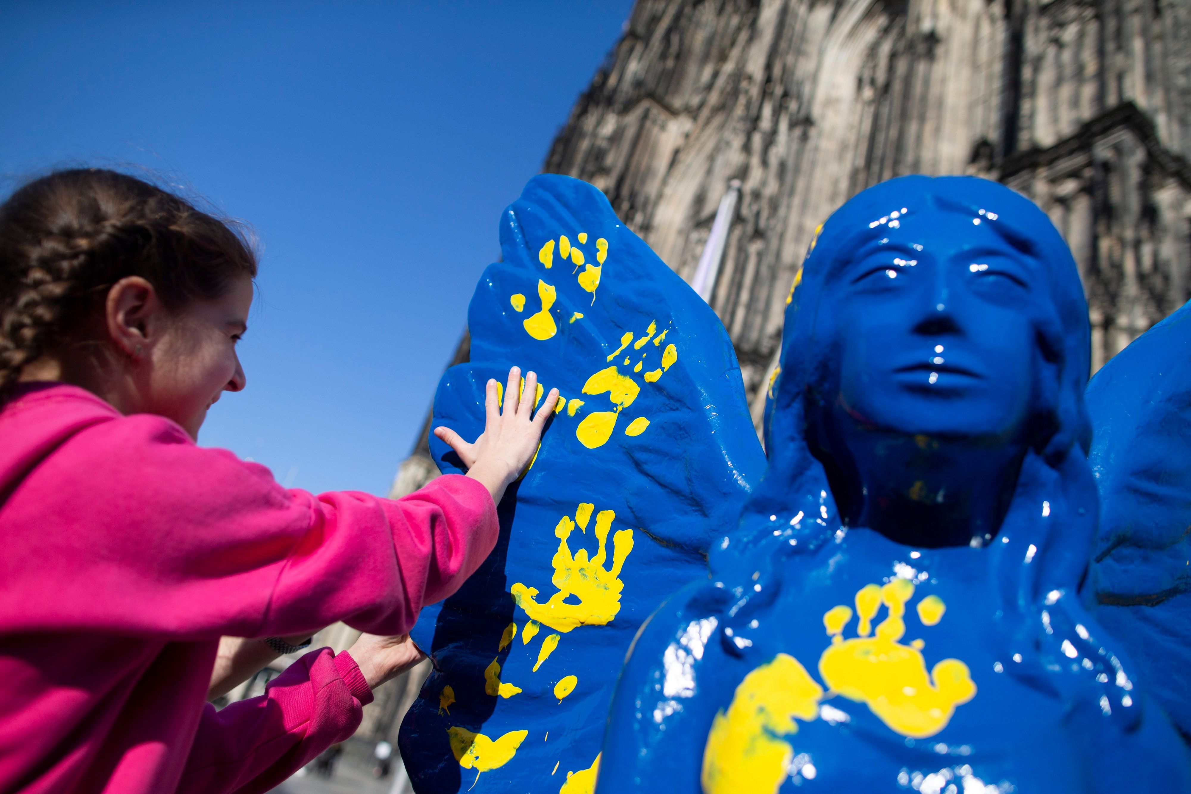 Kinder aus der Ukraine verzieren 2022 mit ihren Händen einen blauen Engel mit gelber Farbe auf der Kölner Domplatte.