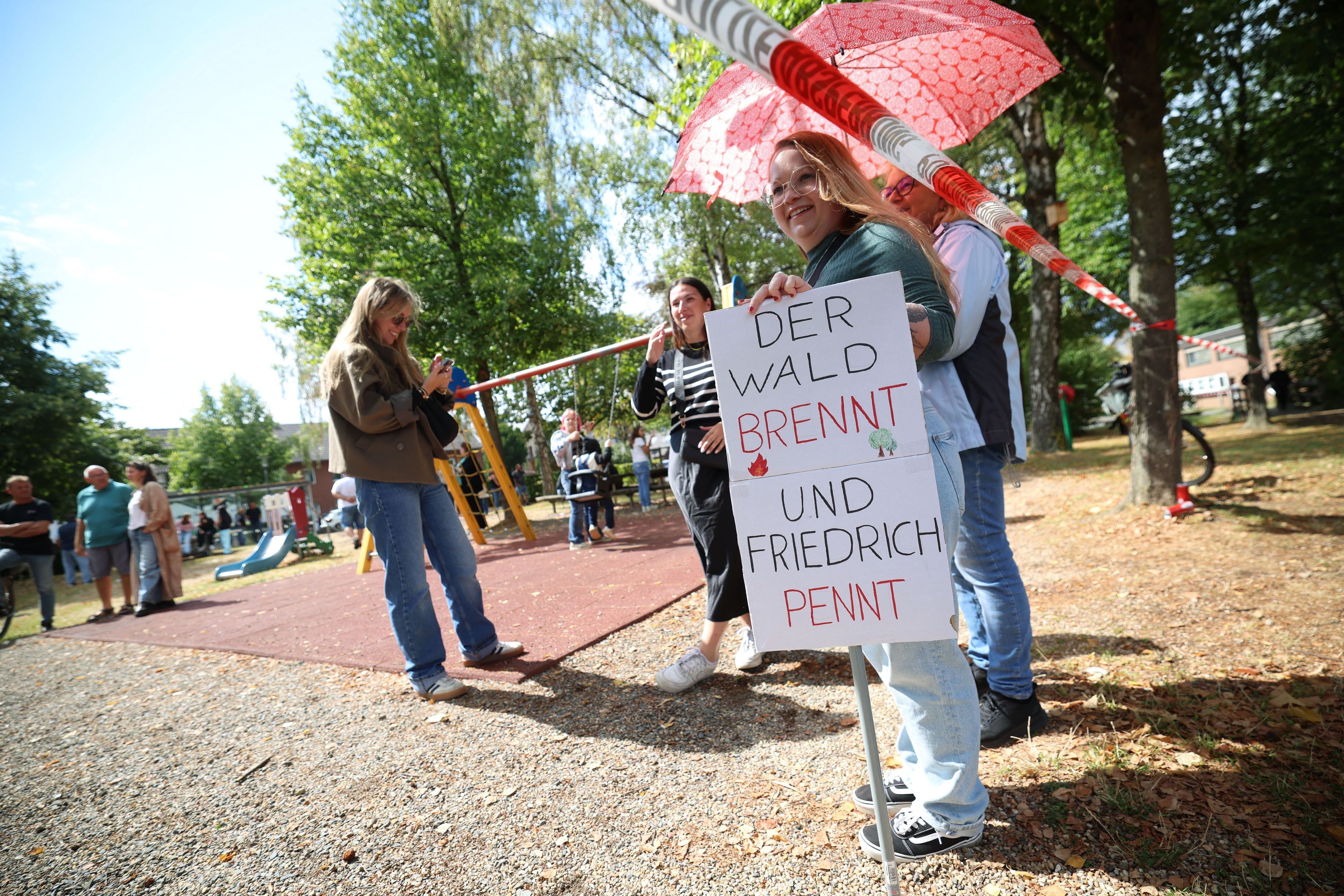 Eine Frau hält in Hürtgenwald ein Schild mit der Aufschrift „Der Wald brennt und Friedrich pennt“ vor dem Besuch von Bundeskanzler Merz in Hürtgenwald-Gey. Zwei Tage nach der vollständigen Löschung des verheerenden Waldbrands in der Eifel besucht Bundeskanzler das Gebiet, um den Helfern zu danken
