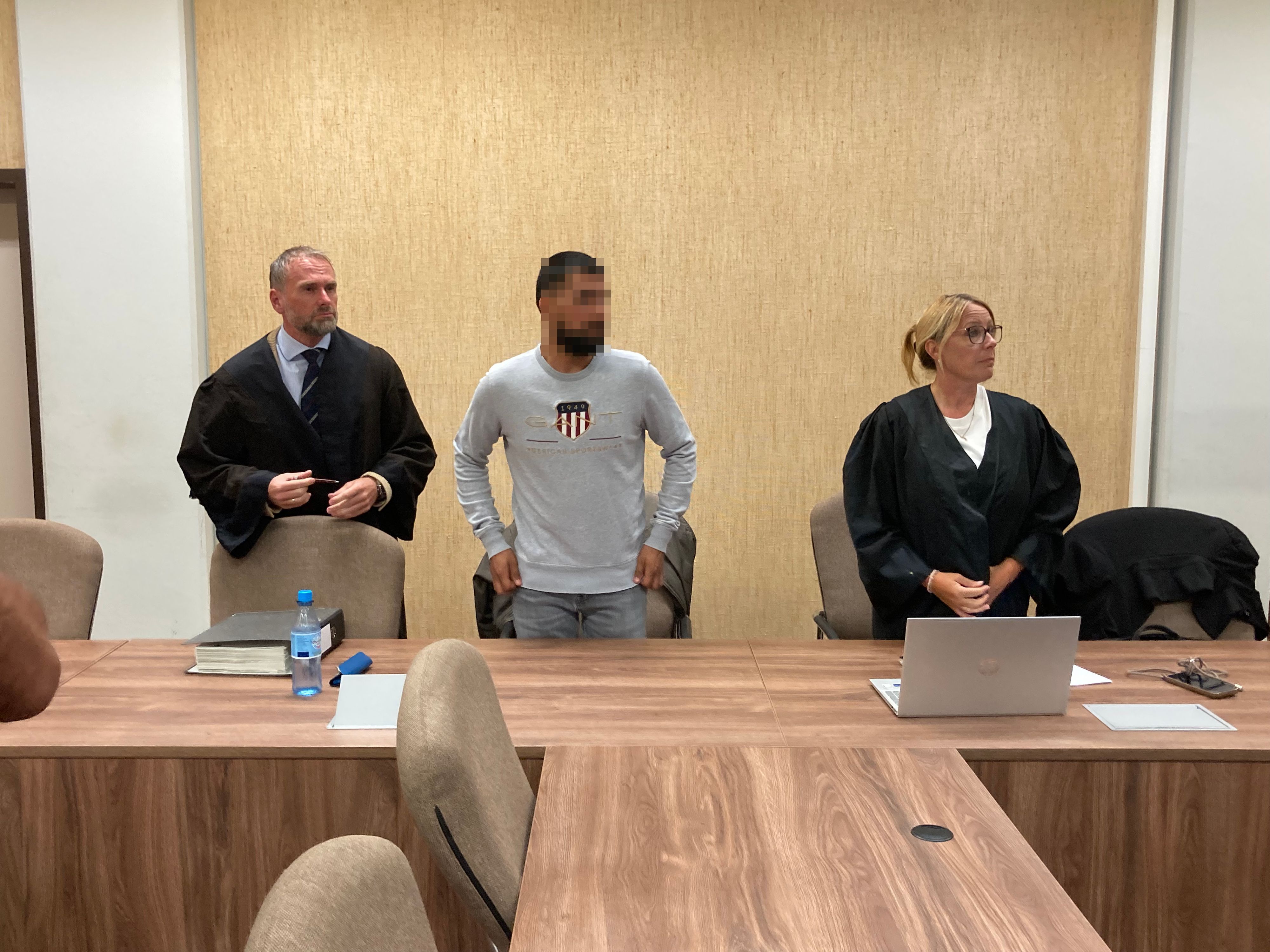 Ein Mann steht im Gericht zwischen seinem Anwalt und seiner Anwältin.