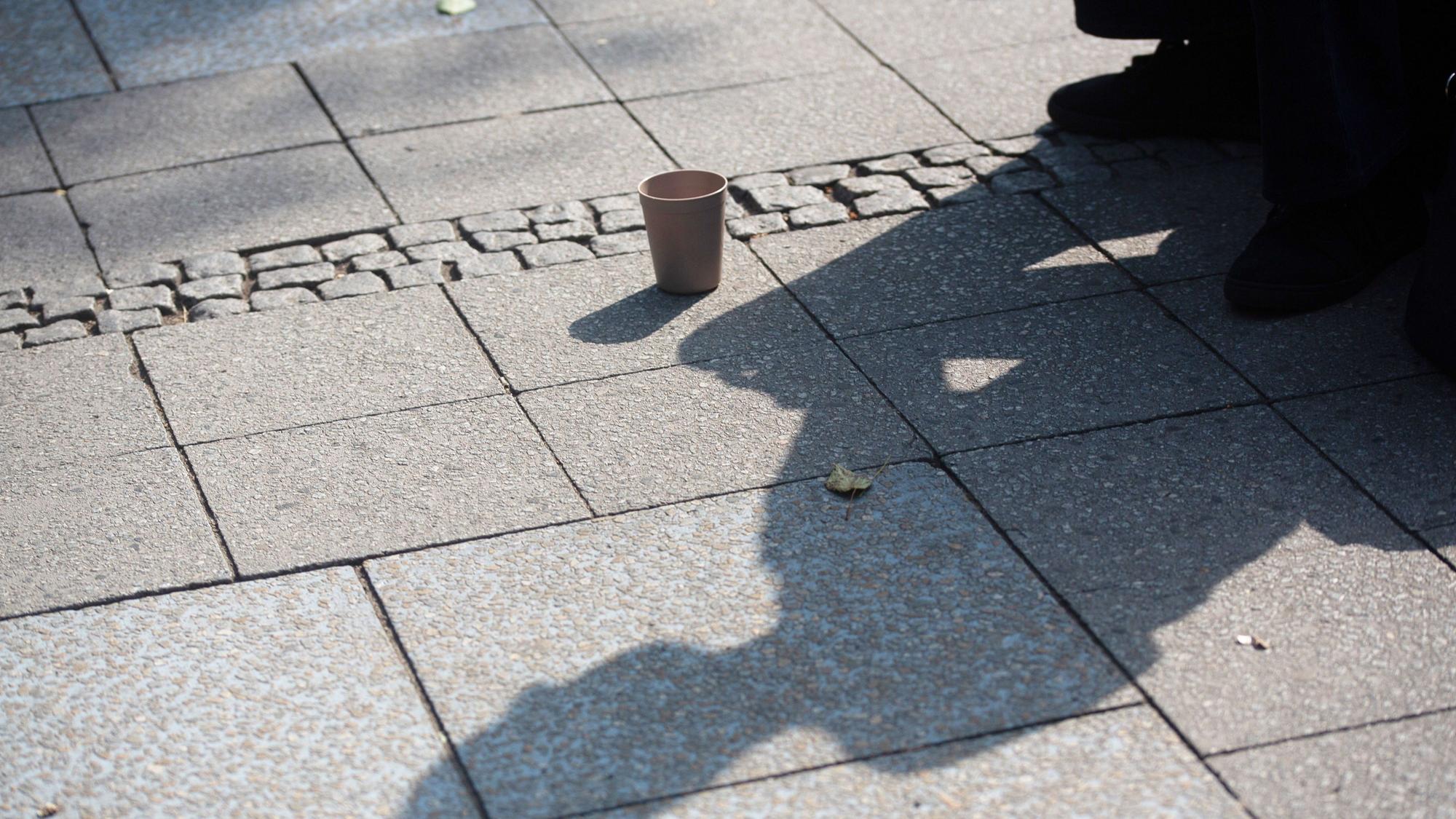 Der Schatten einer Person mit einem Becher für Spenden fällt auf den Gehweg. Im Juli hatte es eine Debatte zu einem möglichen Bettelverbot in Köln gegeben. (Archivfoto)