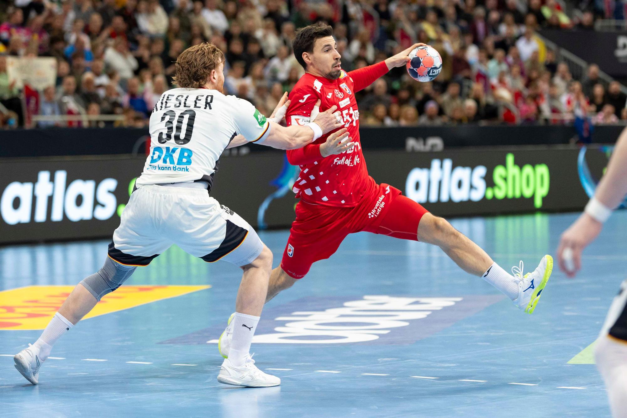 Ein Handballspieler im roten Trikot versucht, sich mit dem Ball gegen einen Gegenspieler im weißen Trikot durchzusetzen. Im Hintergrund ist eine jubelnde Zuschauermenge zu sehen.