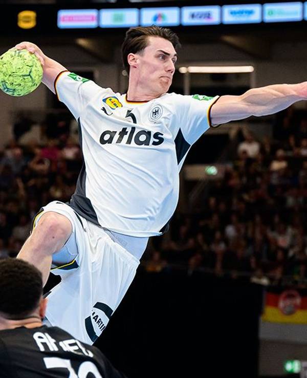 Ein deutscher Handballspieler im weißen Trikot springt zum Wurf, während zwei Gegenspieler ihn zu blocken versuchen.
