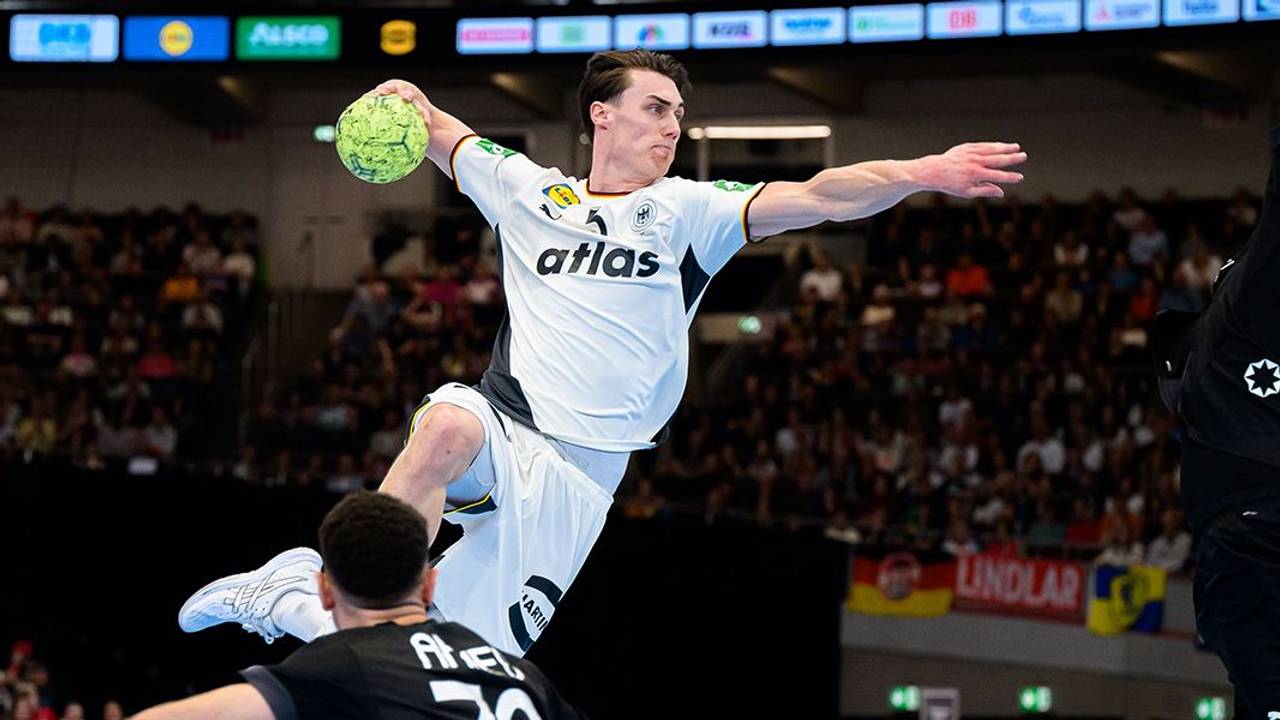 Ein deutscher Handballspieler im weißen Trikot springt zum Wurf, während zwei Gegenspieler ihn zu blocken versuchen.