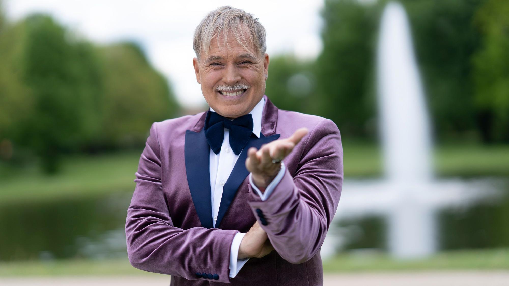 Ein Mann mit grauem Haar und Schnurrbart in einem fliederfarbenen Smoking gestikuliert und schaut in die Kamera