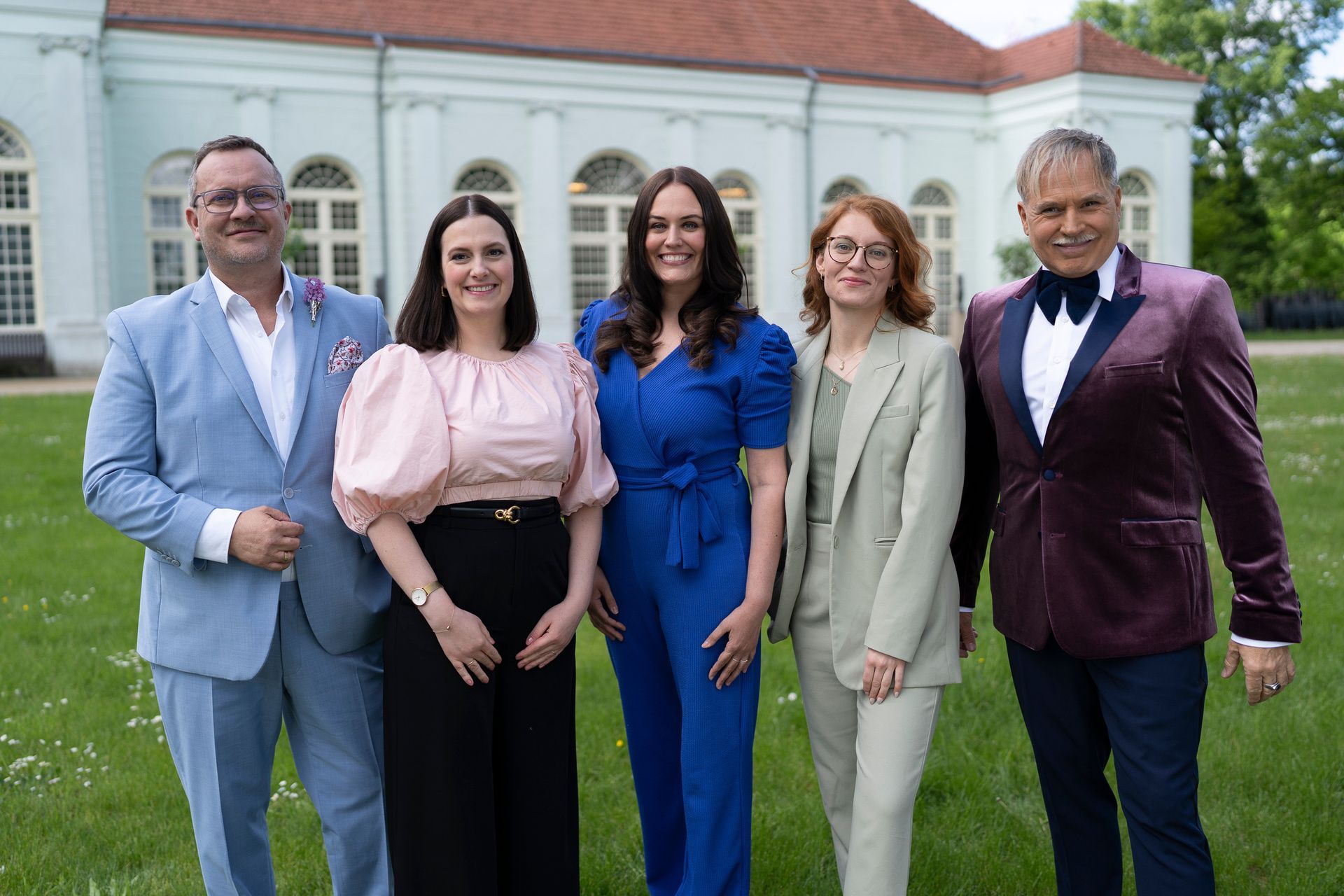 Fünf festlich angezogene Menschen stehen vor einer Orangerie und lachen