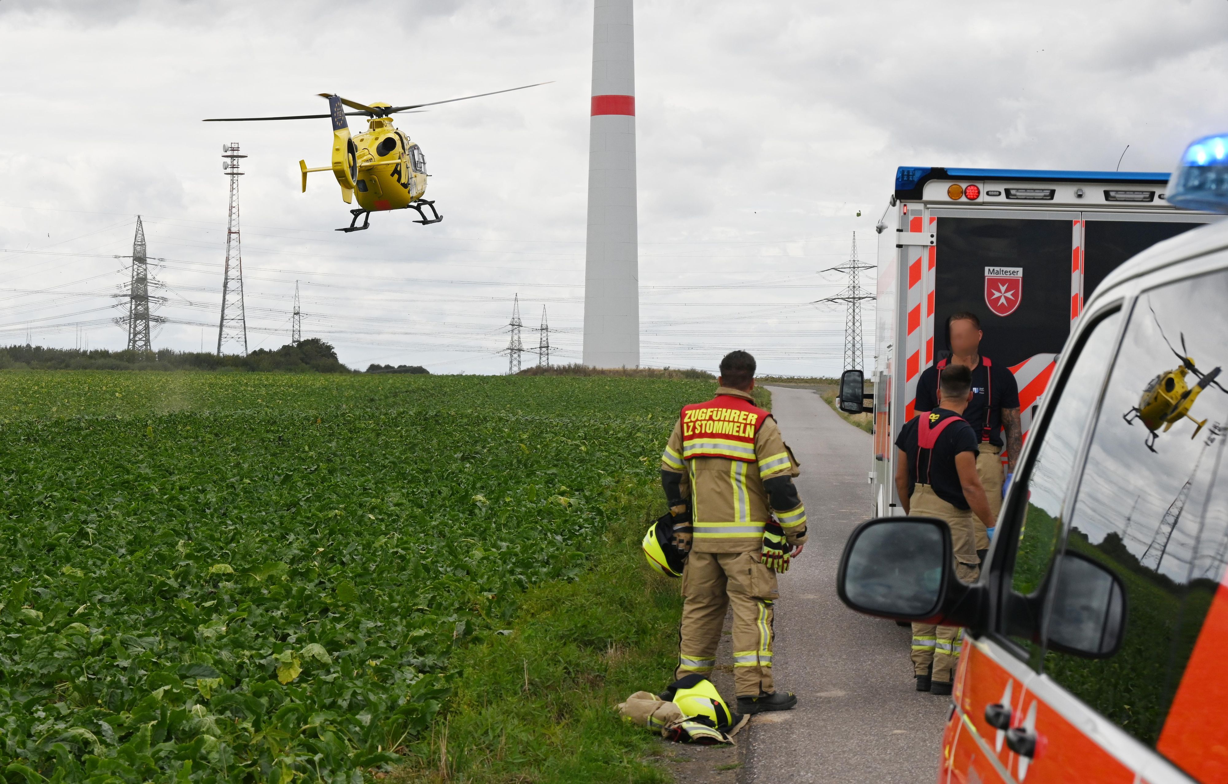 Einsatzfahrzeuge und drei Einsatzkräfte der Feuerwehr stehen an einem Feld, über dem ein Hubschrauber fliegt. Im Hintergrund steht ein Windrad.