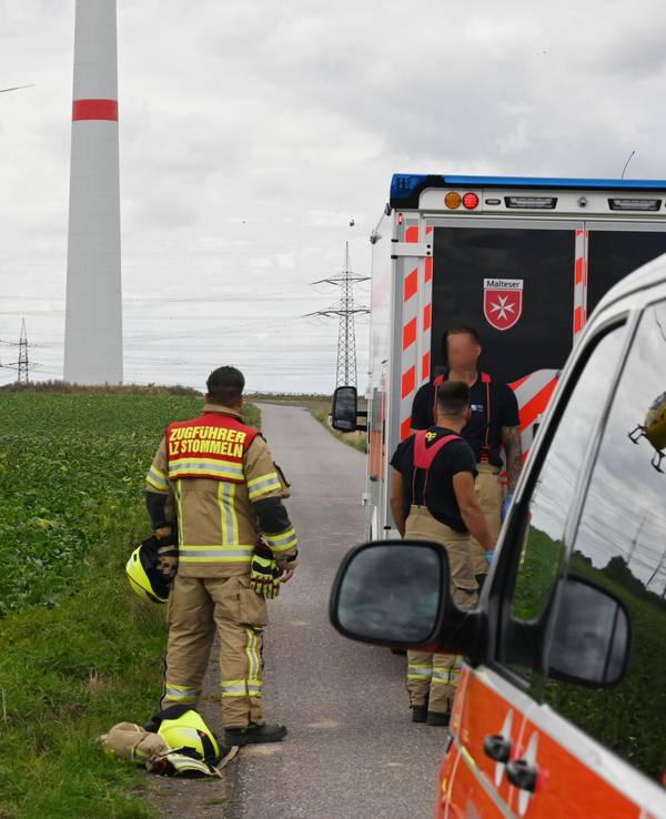 Einsatzfahrzeuge und drei Einsatzkräfte der Feuerwehr stehen an einem Feld, über dem ein Hubschrauber fliegt. Im Hintergrund steht ein Windrad.