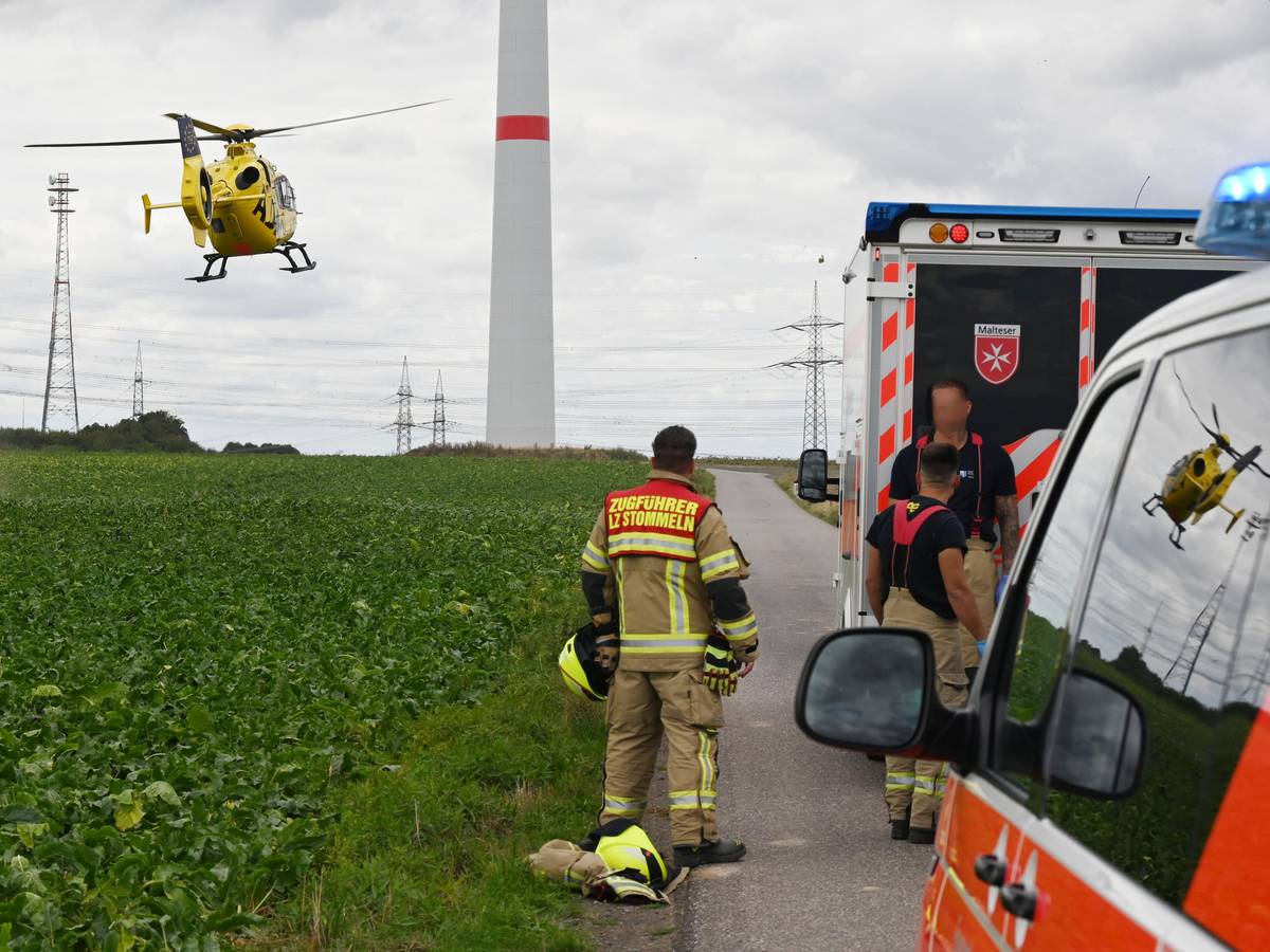 Einsatzfahrzeuge und drei Einsatzkräfte der Feuerwehr stehen an einem Feld, über dem ein Hubschrauber fliegt. Im Hintergrund steht ein Windrad.