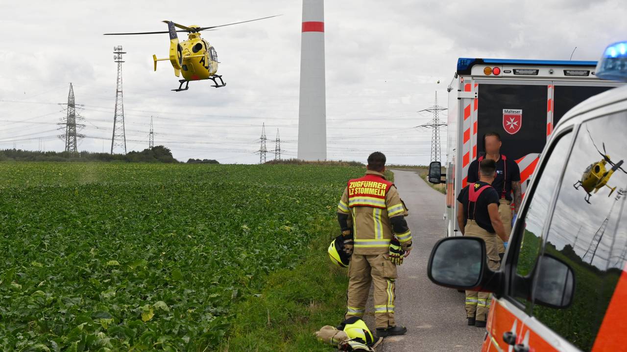 Einsatzfahrzeuge und drei Einsatzkräfte der Feuerwehr stehen an einem Feld, über dem ein Hubschrauber fliegt. Im Hintergrund steht ein Windrad.