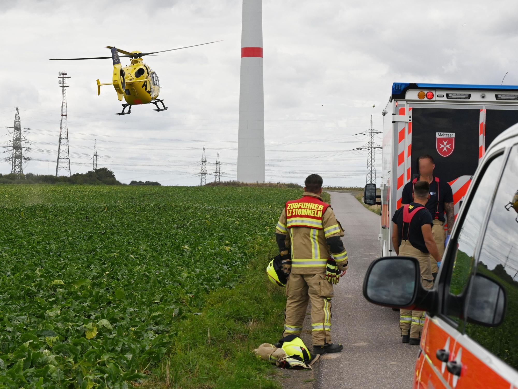 Einsatzfahrzeuge und drei Einsatzkräfte der Feuerwehr stehen an einem Feld, über dem ein Hubschrauber fliegt. Im Hintergrund steht ein Windrad.