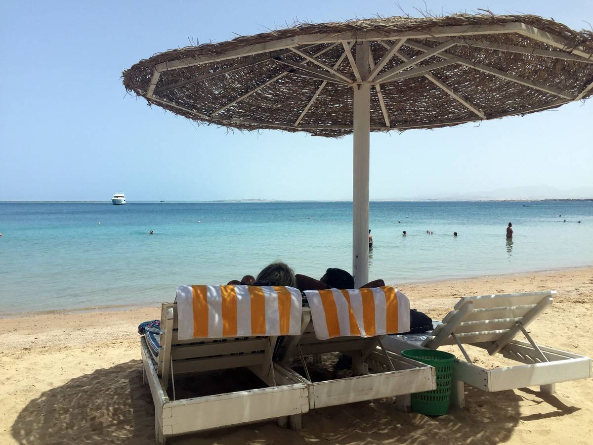 Eine Frau und ein Mann liegen unter einem Sonnenschirm an einem Strand und blicken aufs Meer.