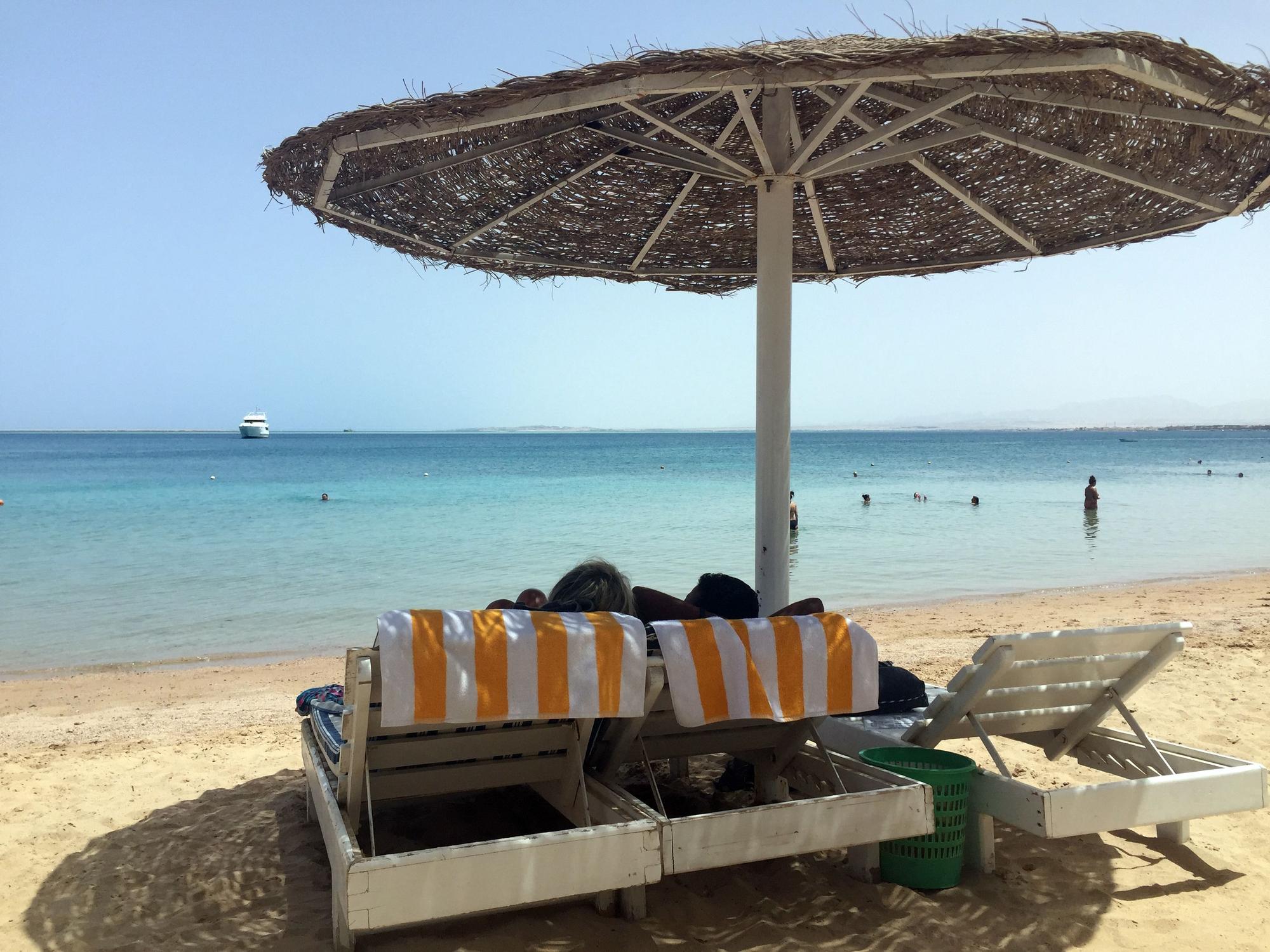 Eine Frau und ein Mann liegen unter einem Sonnenschirm an einem Strand und blicken aufs Meer.