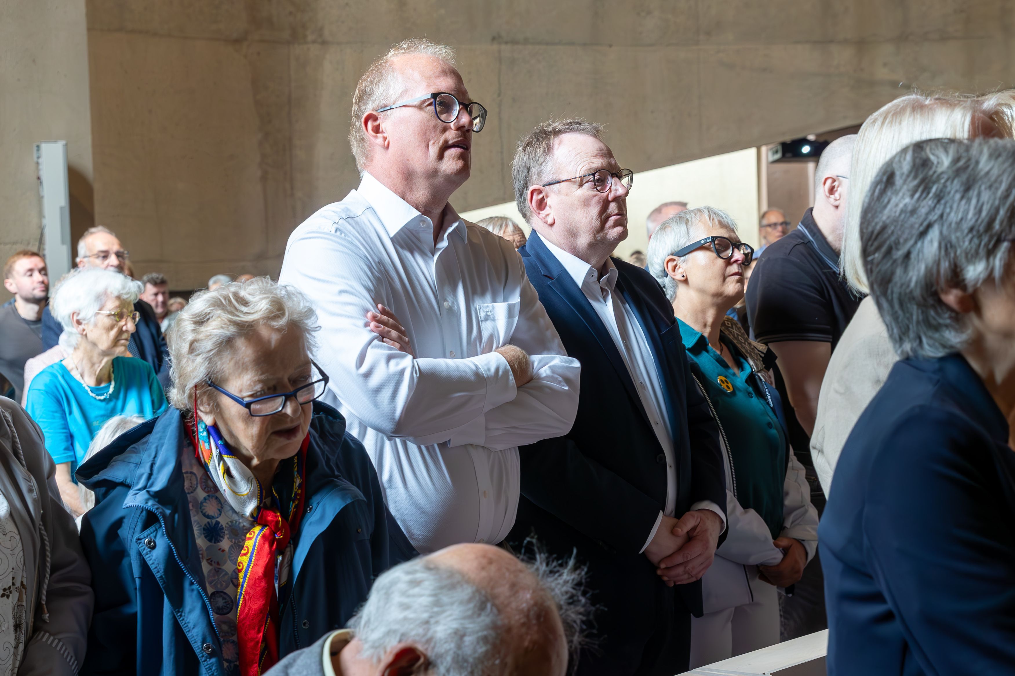 Vollbesetzte Kirchenbänke sind zu sehen. Zwei Männer stehen inmitten der Menge.