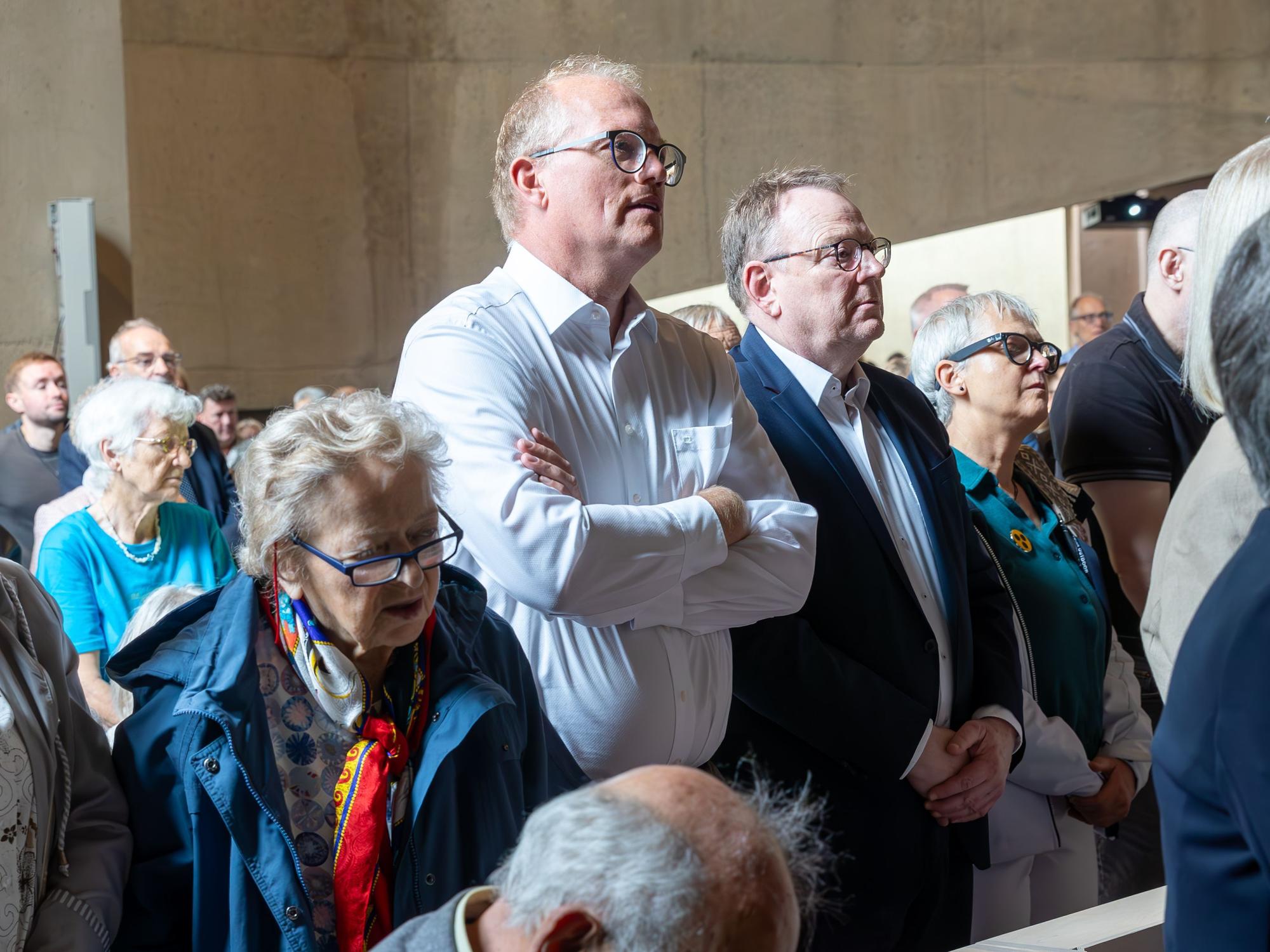 Vollbesetzte Kirchenbänke sind zu sehen. Zwei Männer stehen inmitten der Menge.