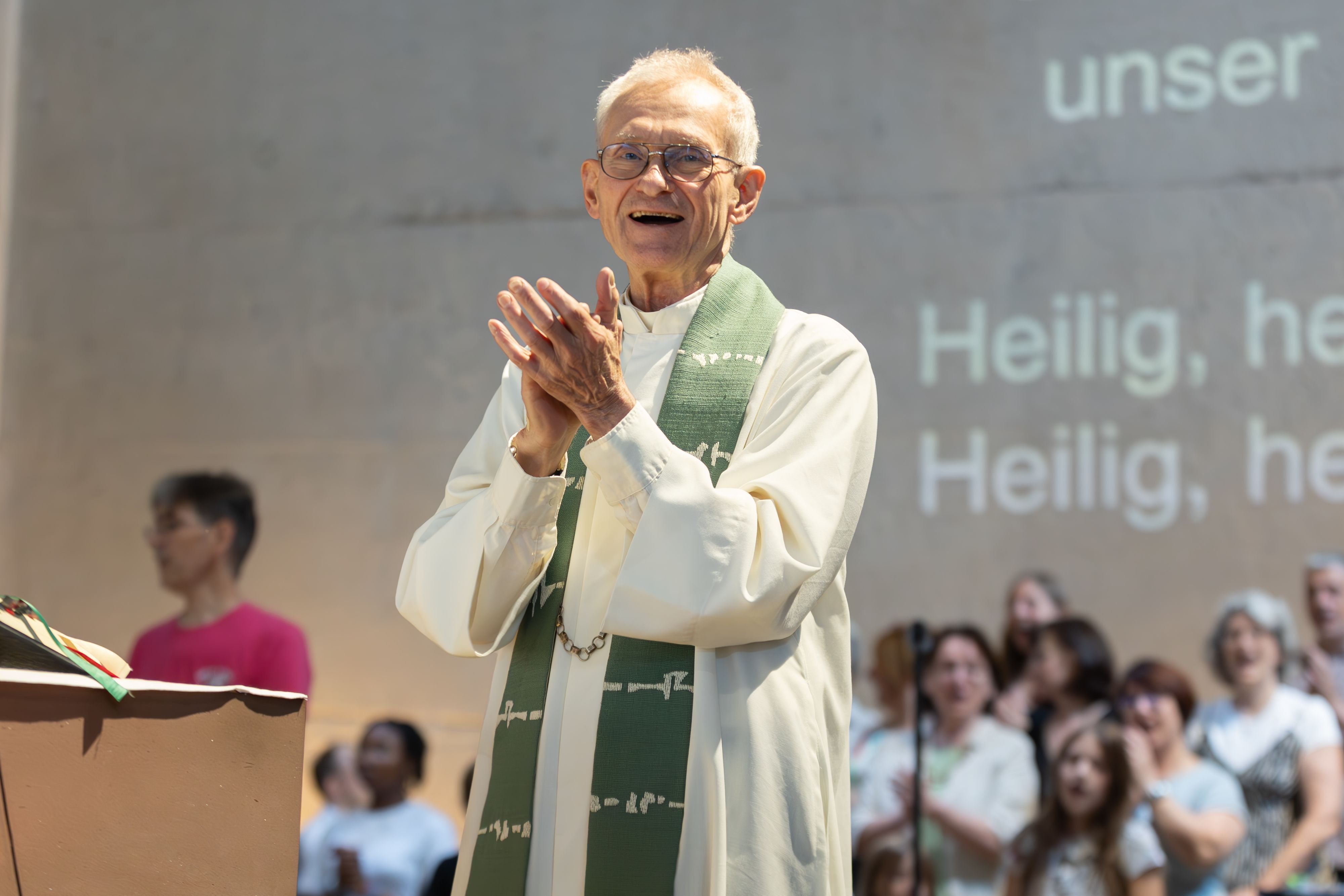 Der Kölner Pfarrer Franz Meurer im Priestergewand klatscht in die Hände. Hinter ihm ist ein Chor zu sehen und Buchstaben an der Wand, die den Text des Liedes zeigen.