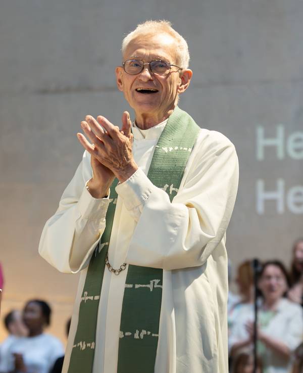 Der Kölner Pfarrer Franz Meurer im Priestergewand klatscht in die Hände. Hinter ihm ist ein Chor zu sehen und Buchstaben an der Wand, die den Text des Liedes zeigen.