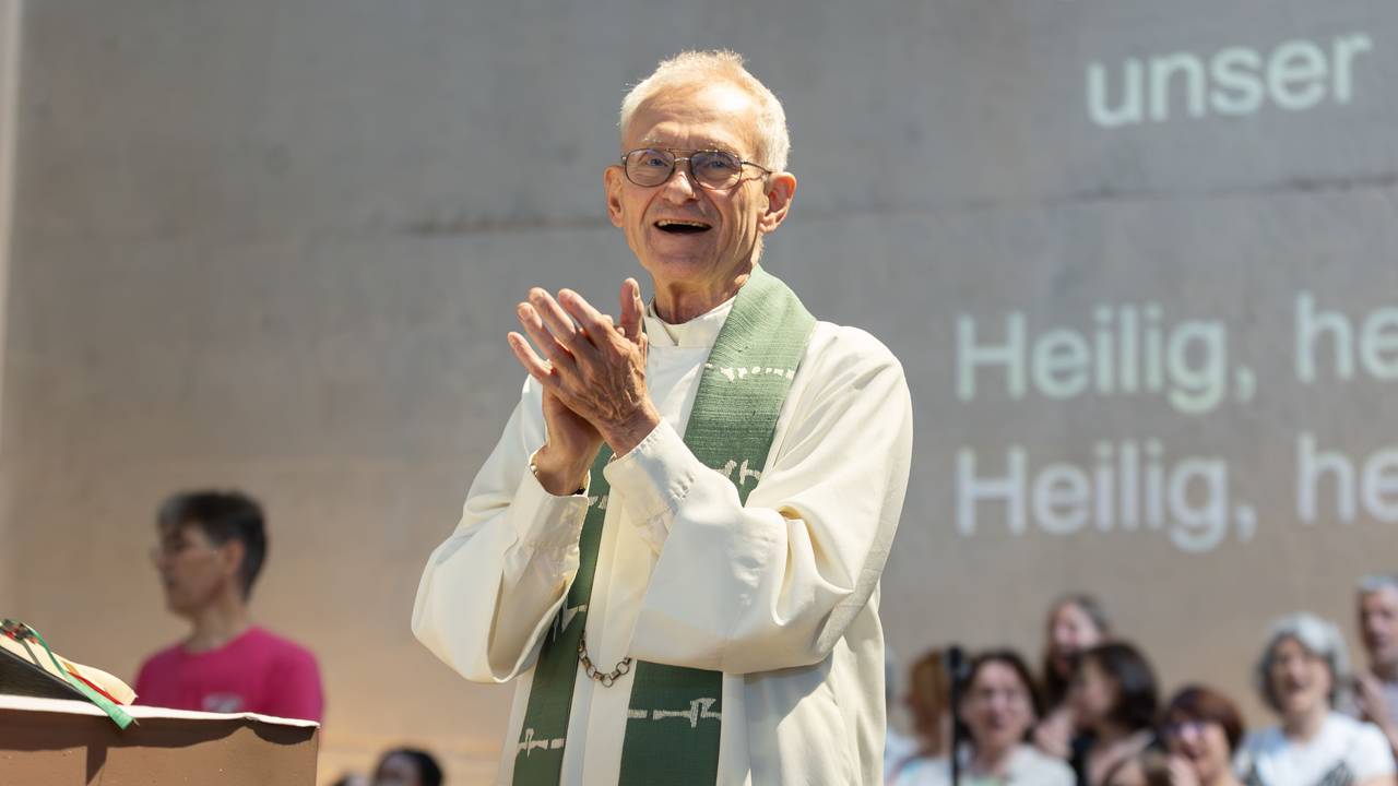 Der Kölner Pfarrer Franz Meurer im Priestergewand klatscht in die Hände. Hinter ihm ist ein Chor zu sehen und Buchstaben an der Wand, die den Text des Liedes zeigen.