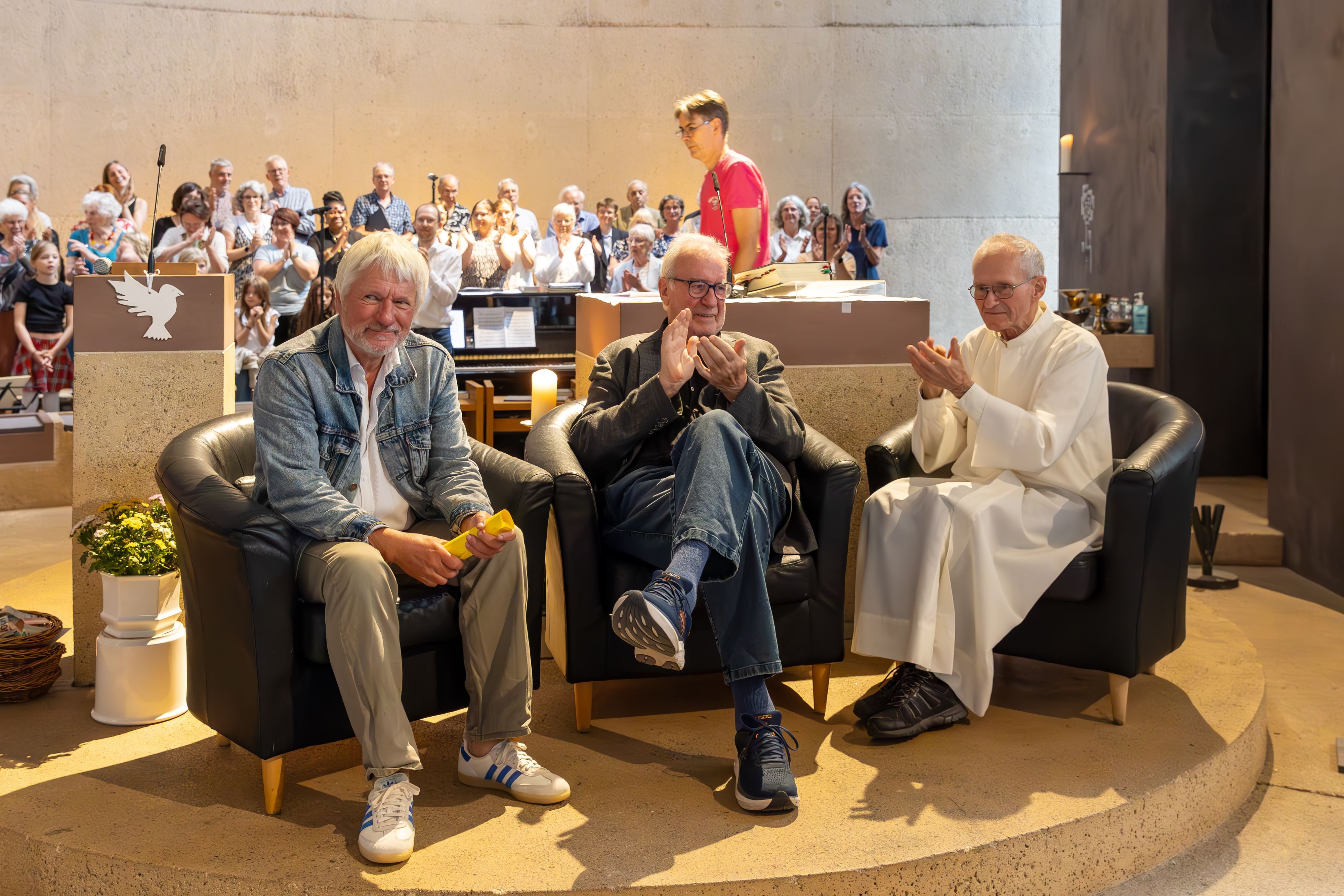 Drei Männer sitzen nebeneinander in Sesseln, hinter ihnen sind Altar, Ambo und der Kirchenchor zu sehen.