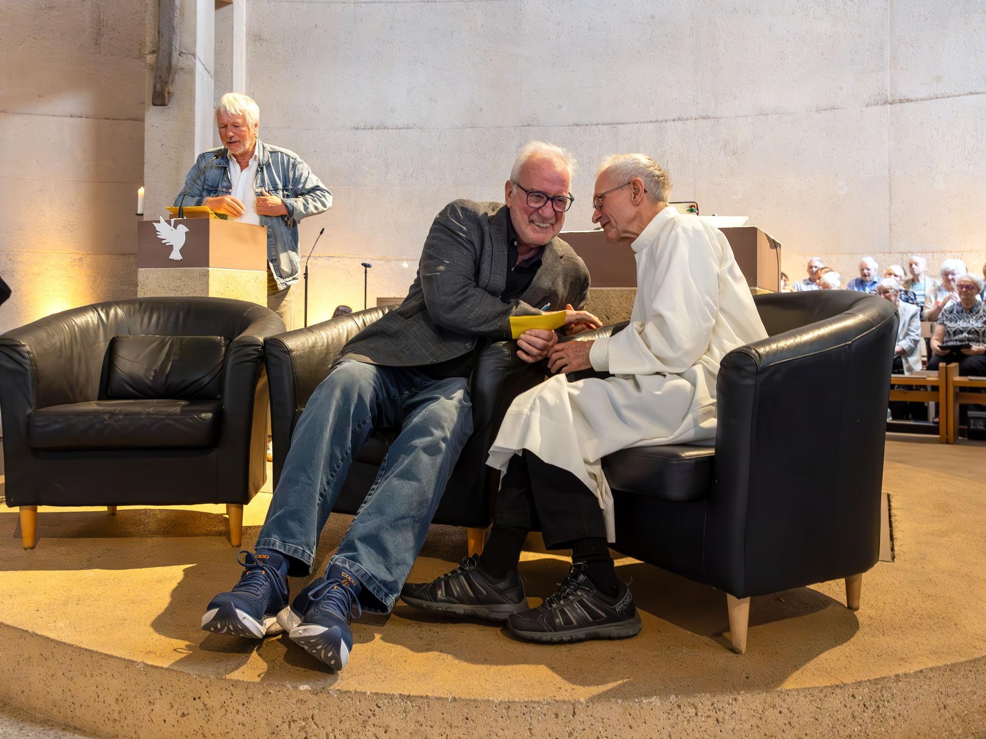 Ein Mann steht am Ambo im Altarraum einer Kirche, auf Sesseln im Vordergrund sitzen zwei Herren, einer trägt die Albe, das Messgewand des Priesters.