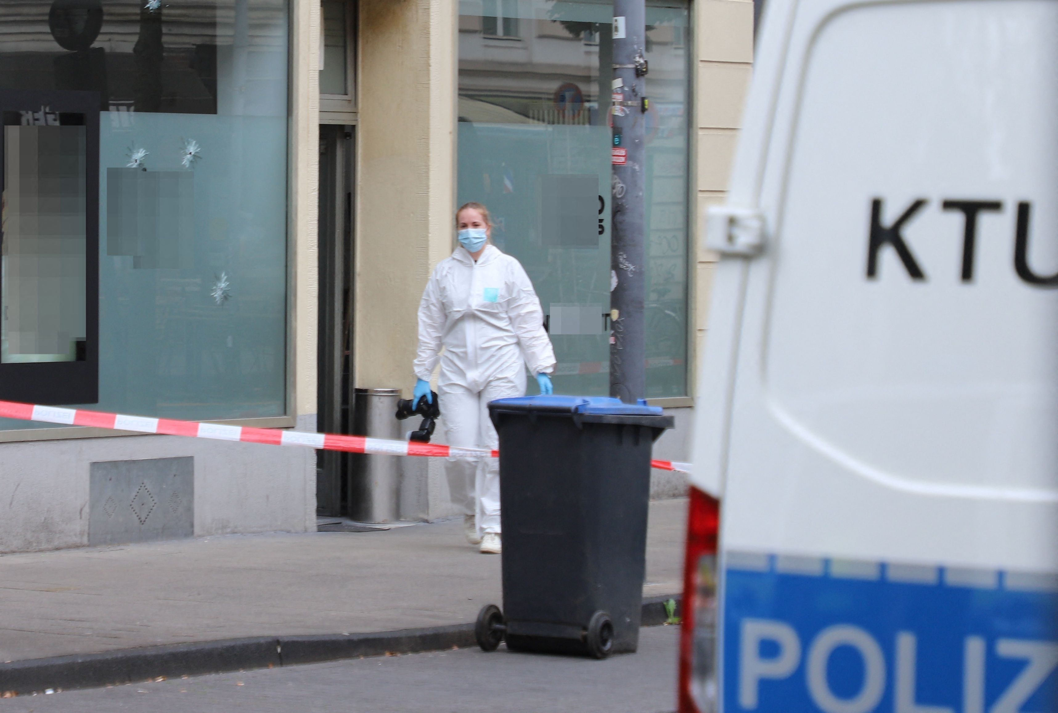 Eine Beamtin in Schutzanzug und mit einer Fotokamera in der Hand ist vor dem Cafè im Einsatz, in dessen Schaufensterscheibe drei Einschusslöcher klaffen. Der Bereich davor ist mit Polizei-Flatterband abgesperrt. Ein Fahrzeug der Spurensicherung parkt davor.