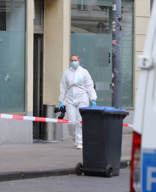 Eine Beamtin in Schutzanzug und mit einer Fotokamera in der Hand ist vor dem Cafè im Einsatz, in dessen Schaufensterscheibe drei Einschusslöcher klaffen. Der Bereich davor ist mit Polizei-Flatterband abgesperrt. Ein Fahrzeug der Spurensicherung parkt davor.