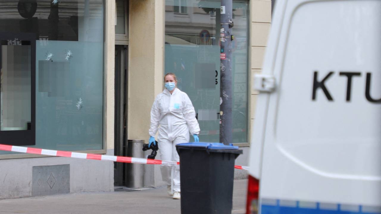 Eine Beamtin in Schutzanzug und mit einer Fotokamera in der Hand ist vor dem Cafè im Einsatz, in dessen Schaufensterscheibe drei Einschusslöcher klaffen. Der Bereich davor ist mit Polizei-Flatterband abgesperrt. Ein Fahrzeug der Spurensicherung parkt davor.
