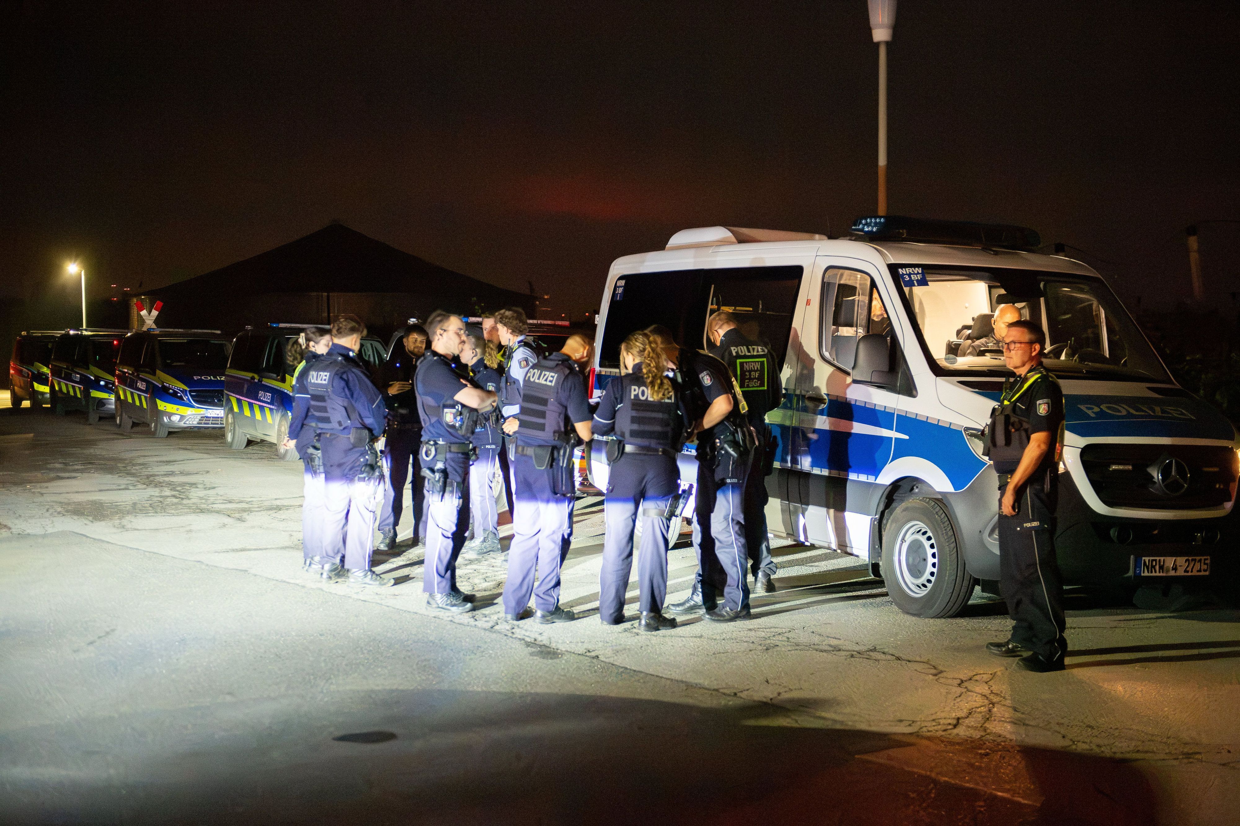 Mehrere Polizeikräfte stehen vor einem Polizeifahrzeug, dahinter stehen weitere Wagen der Polizei.