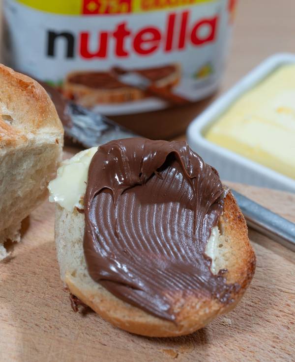 Brötchen mit Nuss-Nougat-Aufstrich und Nutella-Glas im Hintergrund.