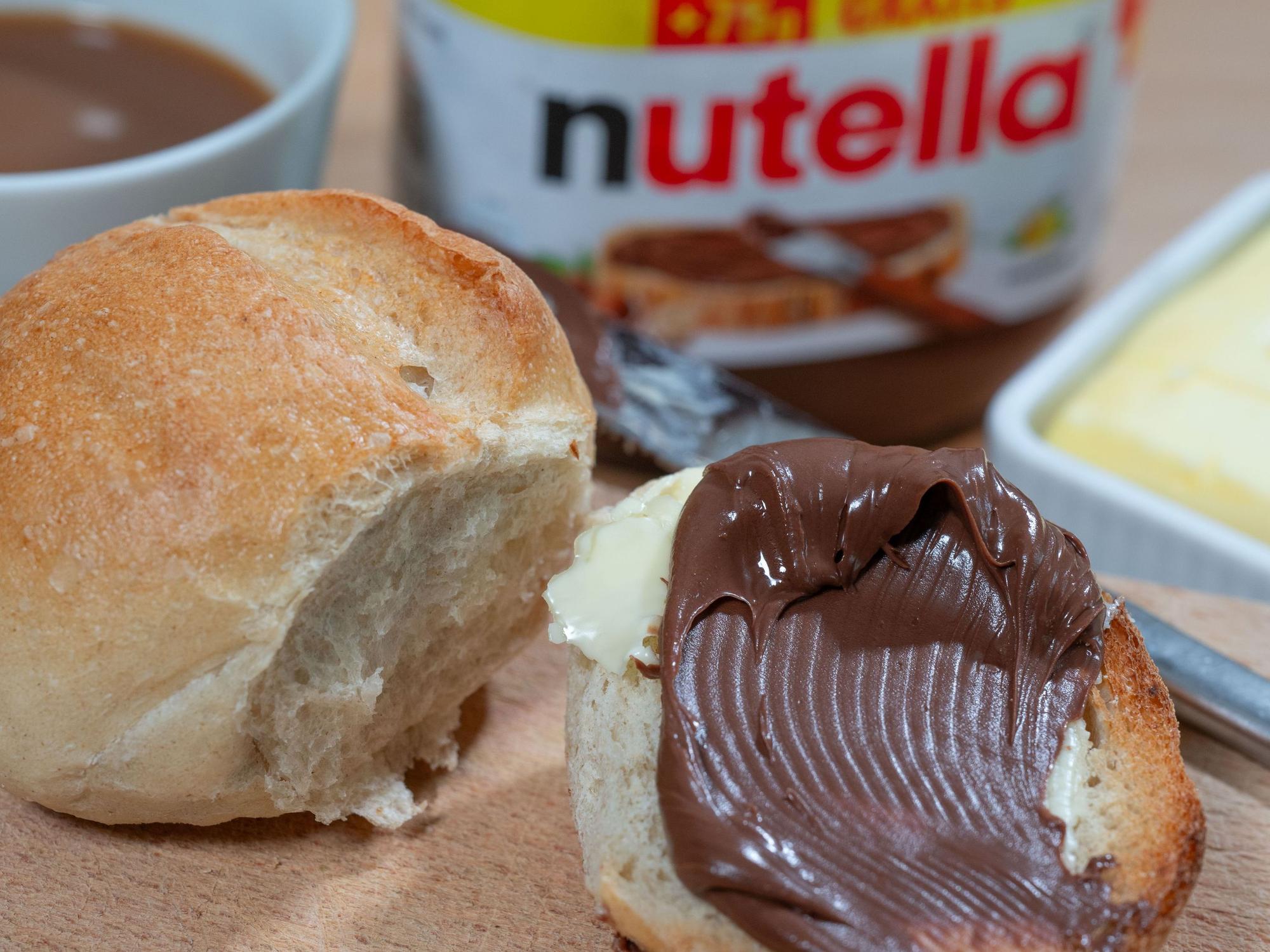 Brötchen mit Nuss-Nougat-Aufstrich und Nutella-Glas im Hintergrund.