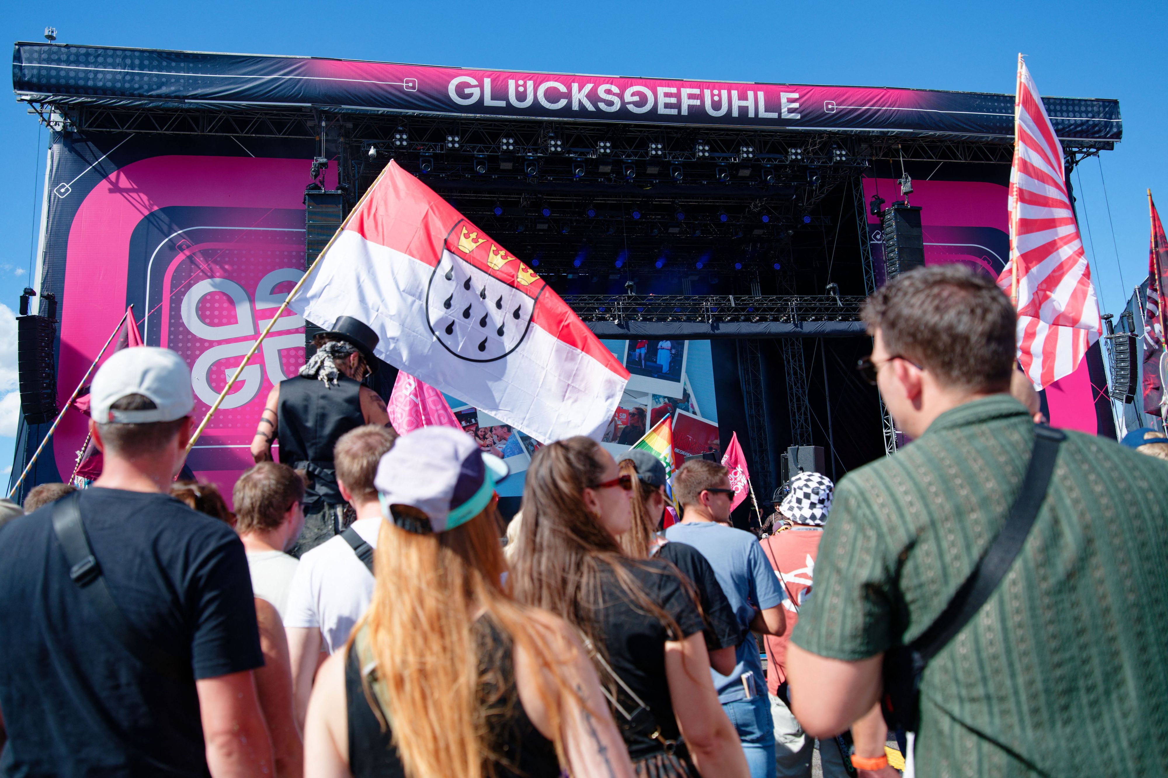 Besucher stehen vor einer Bühne beim Glücksgefühle-Festival auf dem Hockenheimring und halten eine Kölner Stadtflagge. Auch der Kölner Männerchor „Die Gürngürtelrosen“ trat auf dem Hockenheimring auf.