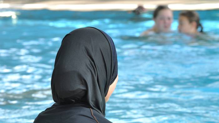 Eine muslimische Schülerin sitzt in einem Ganzkörperbadeanzug (Burkini) am Rande eines Schwimmbeckens.