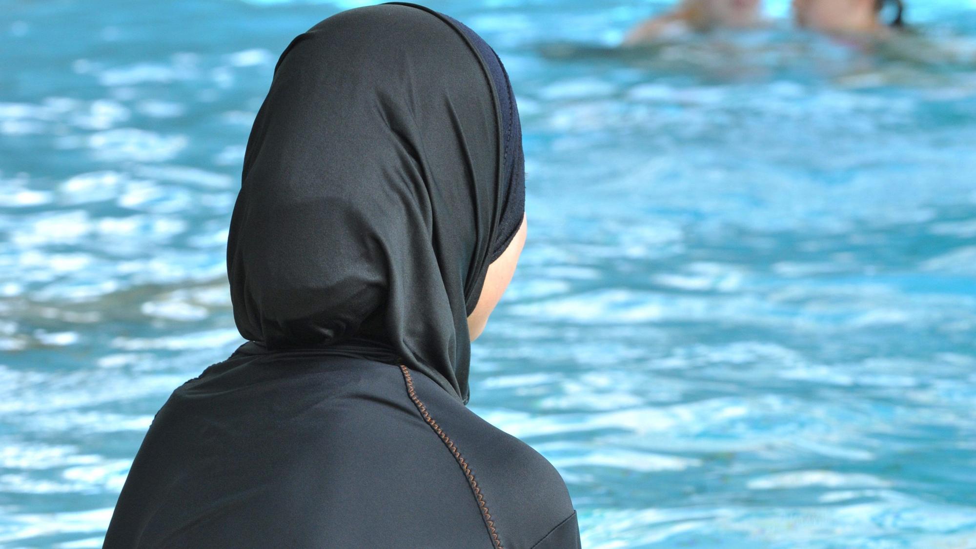 Eine muslimische Schülerin sitzt in einem Ganzkörperbadeanzug (Burkini) am Rande eines Schwimmbeckens.