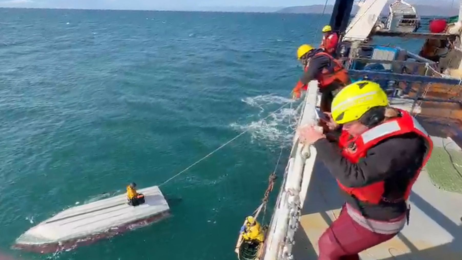 Ein Junge hockt auf einem gekenterten Boot und wird von einer Schiffsbesatzung gerettet.