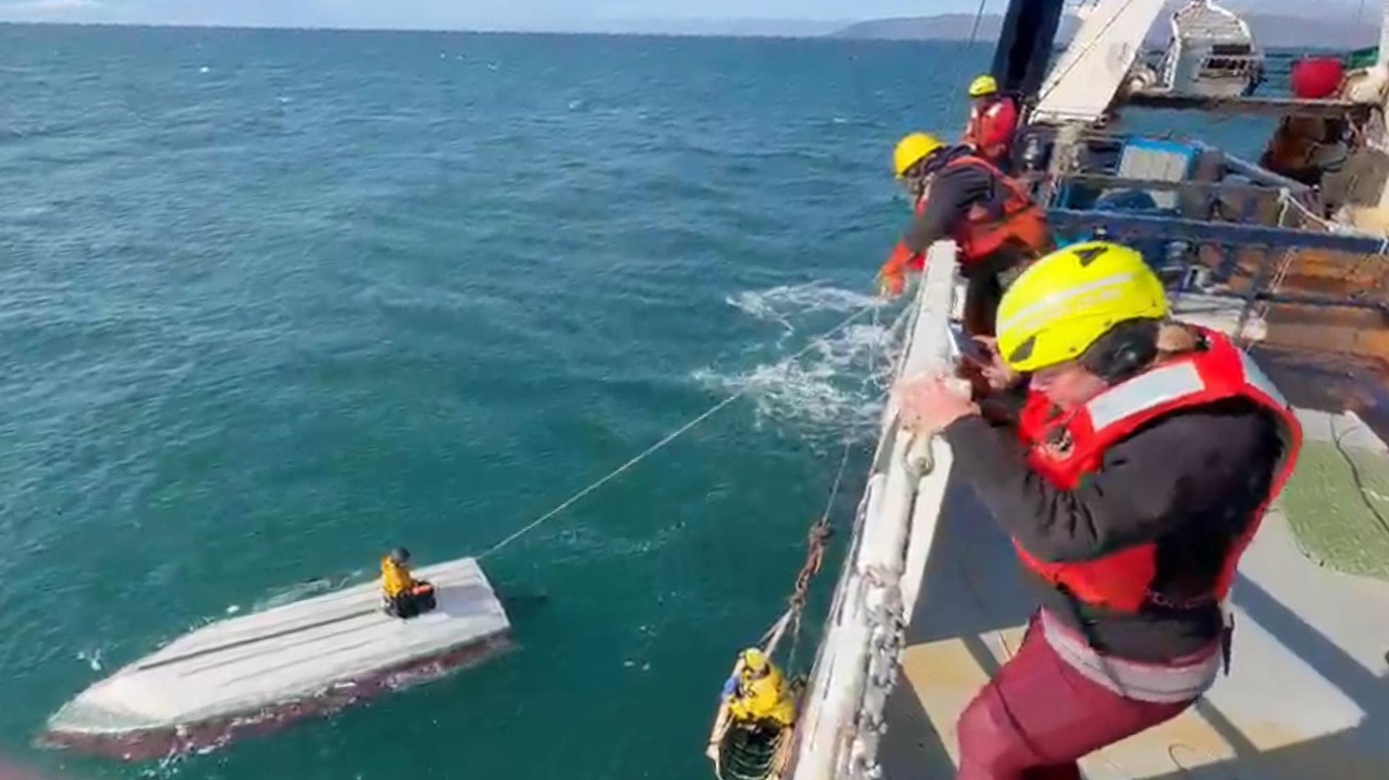Ein Junge hockt auf einem gekenterten Boot und wird von einer Schiffsbesatzung gerettet.