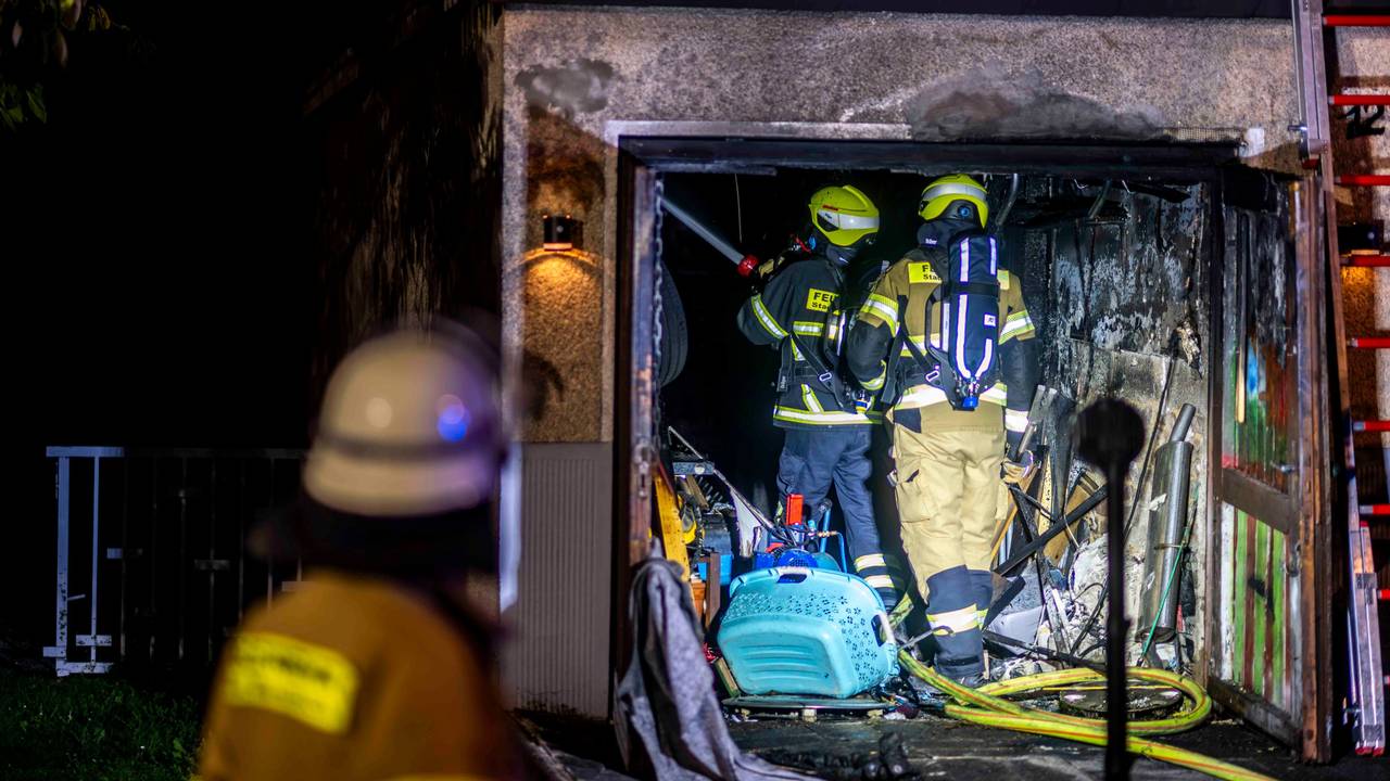Zwei Feuerwehrleute stehen in der verrußten Garage, links ist im Vordergrund ein Feuerwehrmann von hinten unscharf zu sehen.