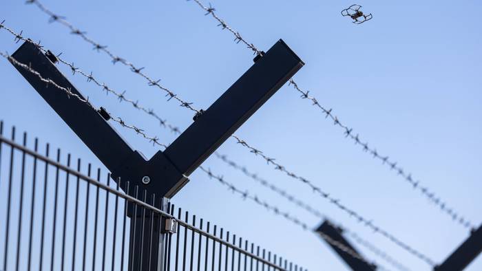Fliegende Drohne am Horizont hinter einem Sicherheitszaun mit Stacheldraht.