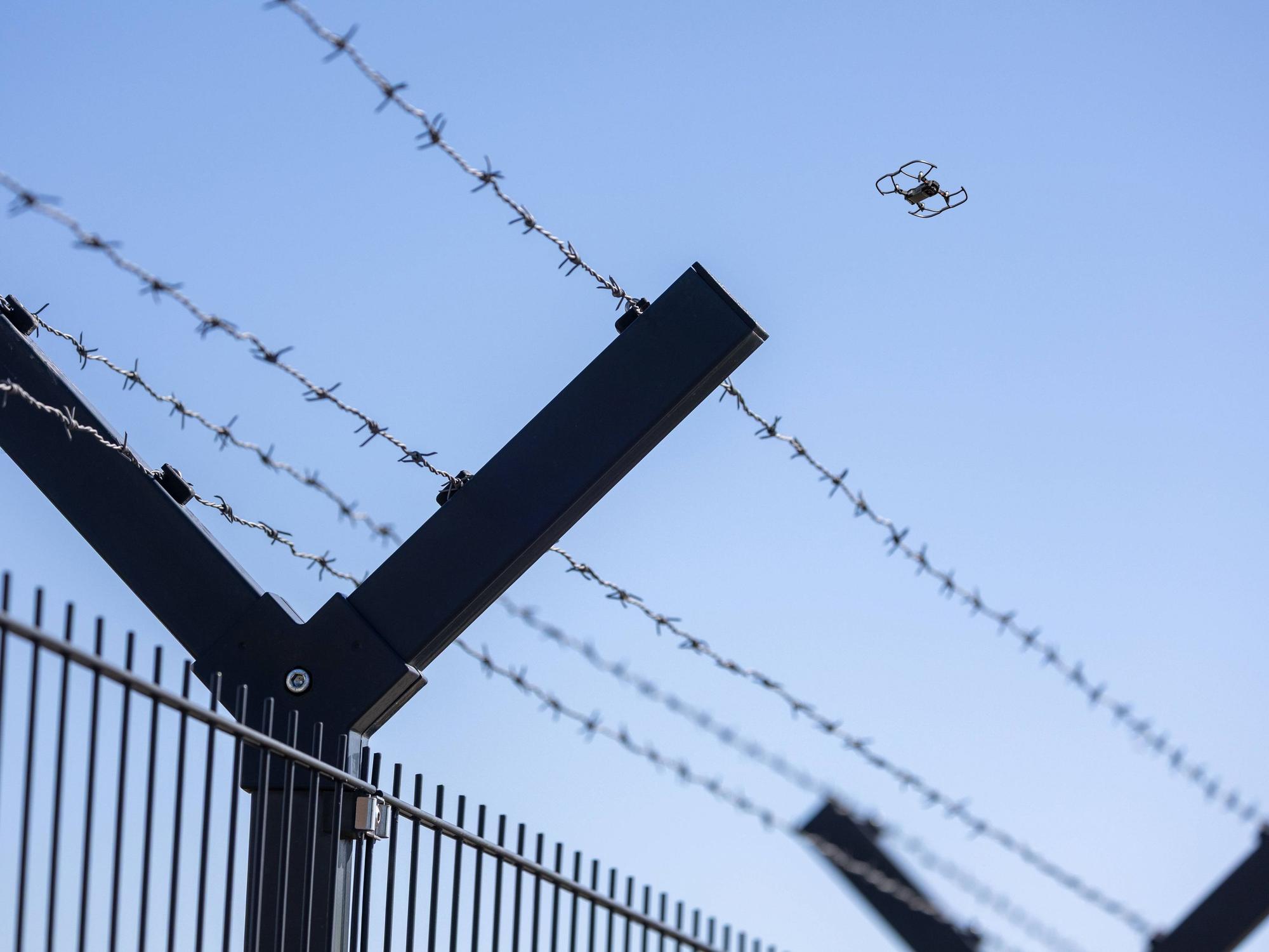 Fliegende Drohne am Horizont hinter einem Sicherheitszaun mit Stacheldraht.
